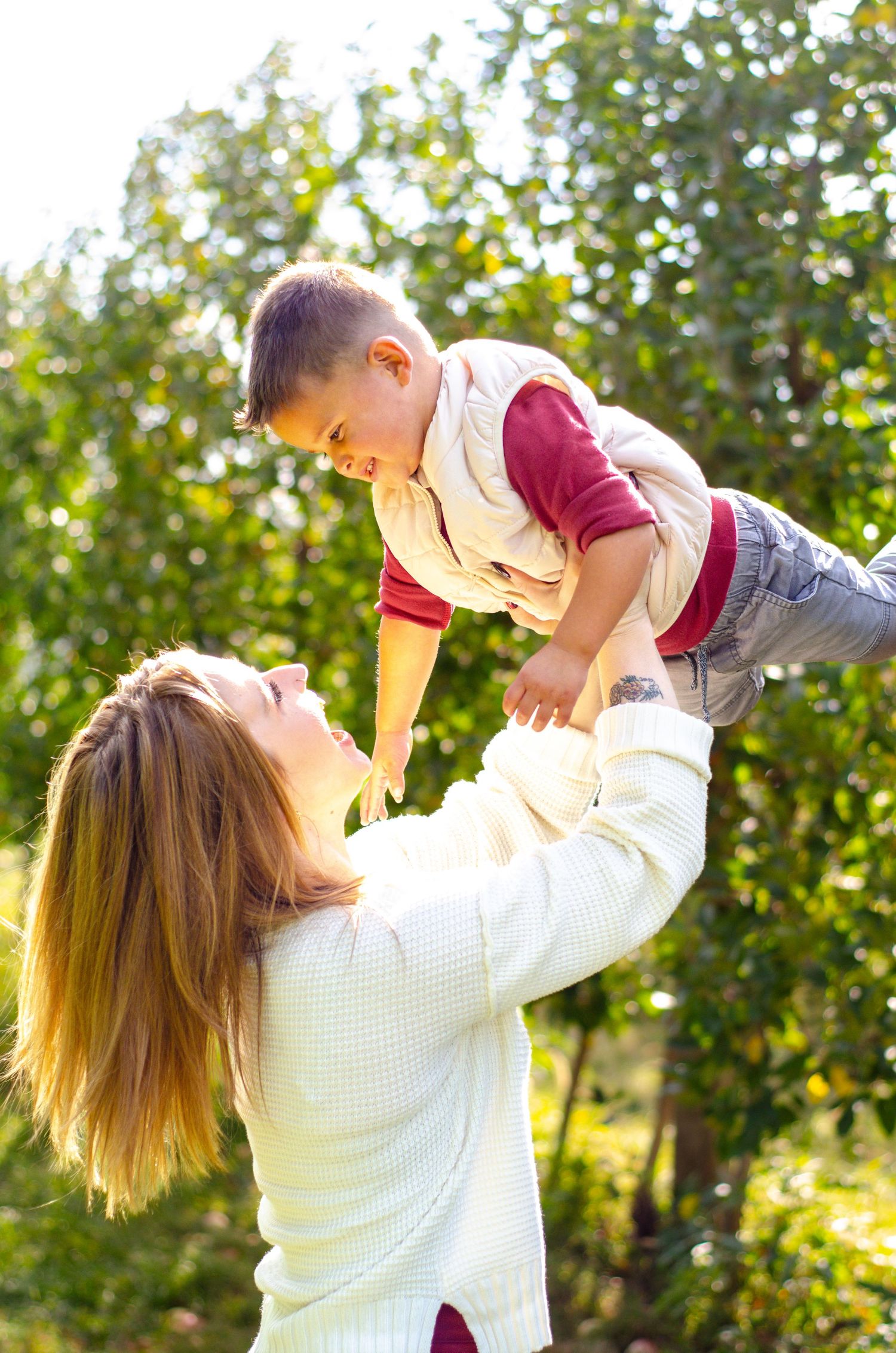 Joyful outdoor scene with a small child being lifted up into the air against sunlit trees. Family photos by KM Conceptions Photography.