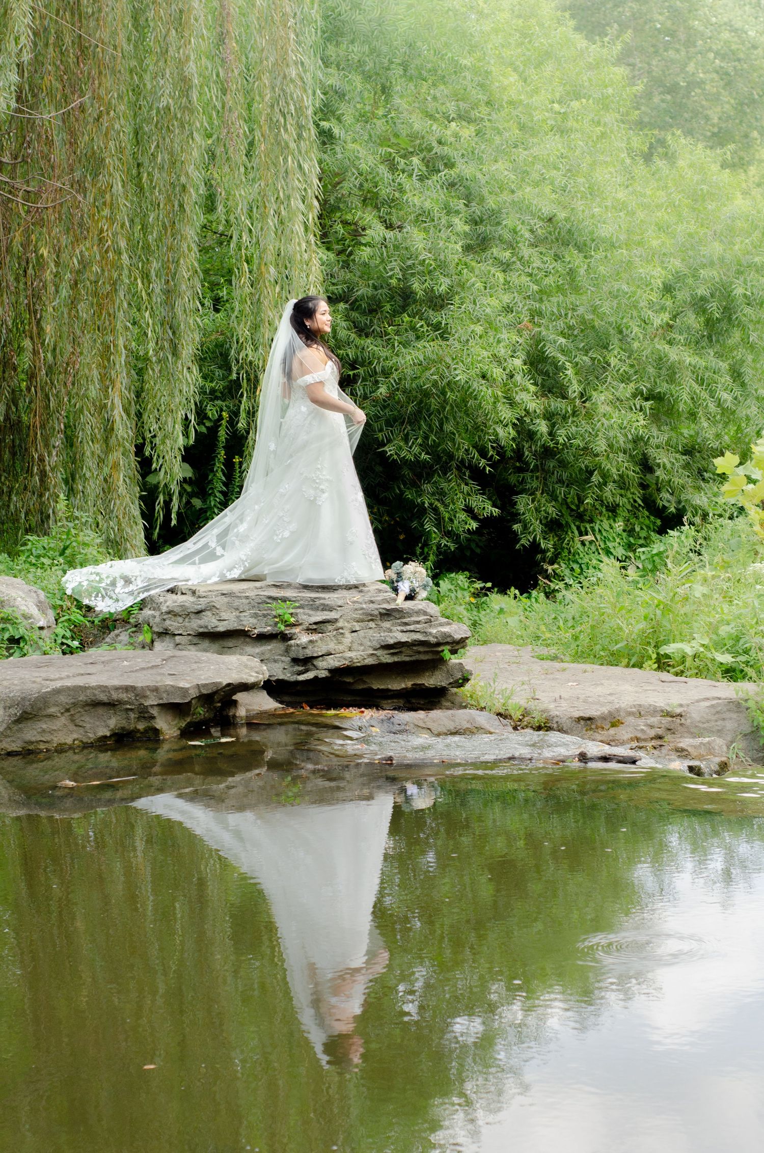 A white wedding dress reflects beautifully in a serene pond surrounded by lush green forest scenery.