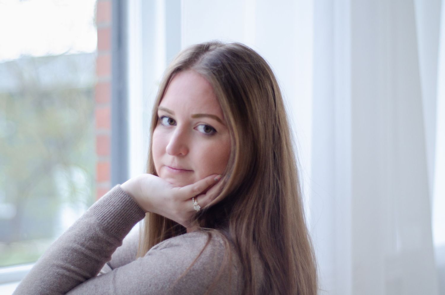 Young woman with long blonde hair resting chin on hand, gazing thoughtfully near a bright window.