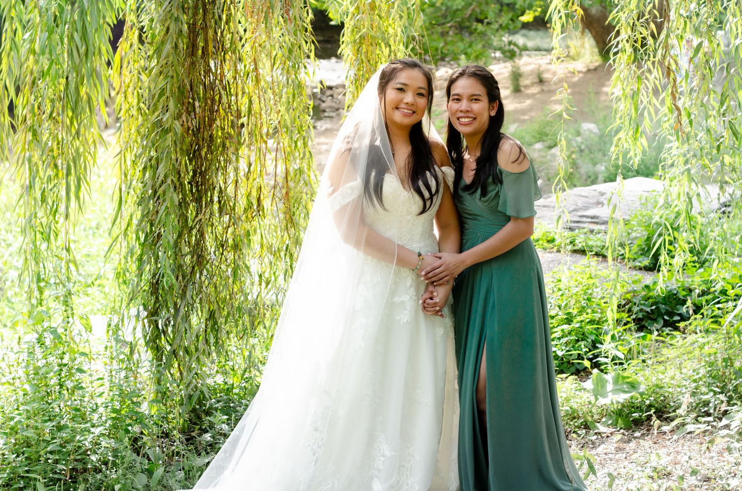 A bride in white and bridesmaid in sage green pose together beneath drooping willow branches.