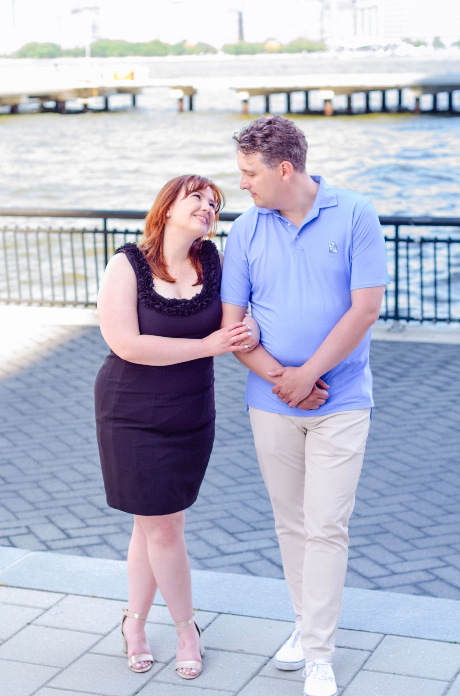 Romantic couple in blue and black outfits embracing by waterfront railing.  Engagement photos by KM Conceptions Photography in New York.
