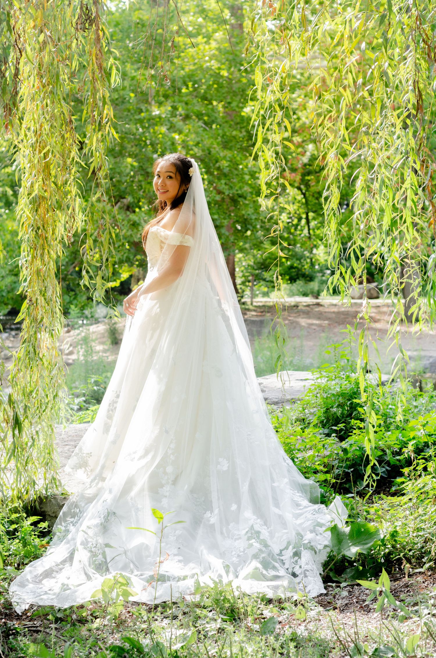 A flowing white wedding gown with delicate lace details is photographed in a garden setting under drooping willow branches.