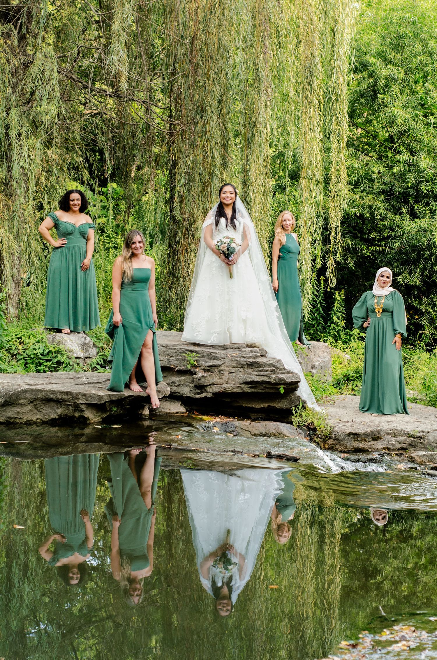 Wedding party in green dresses poses on large rock formation beside reflective pond in natural outdoor setting.