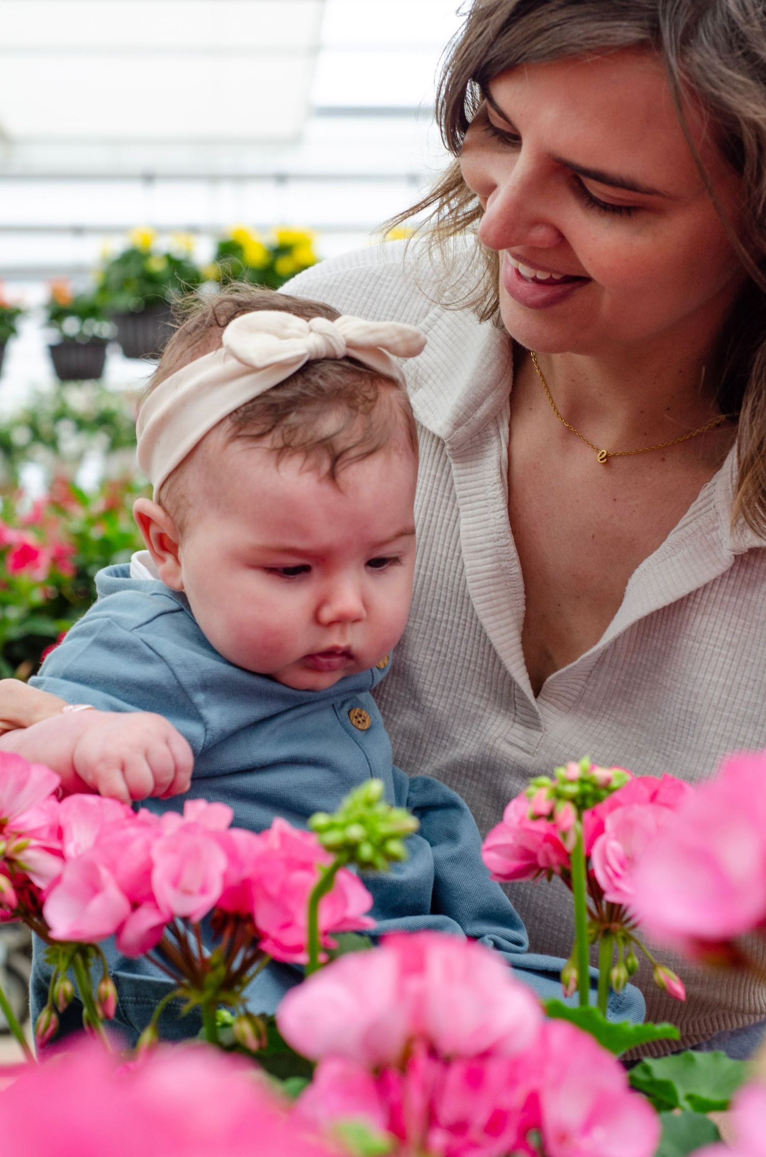 A baby in a blue outfit leans over to smell bright pink flowers in a garden setting.