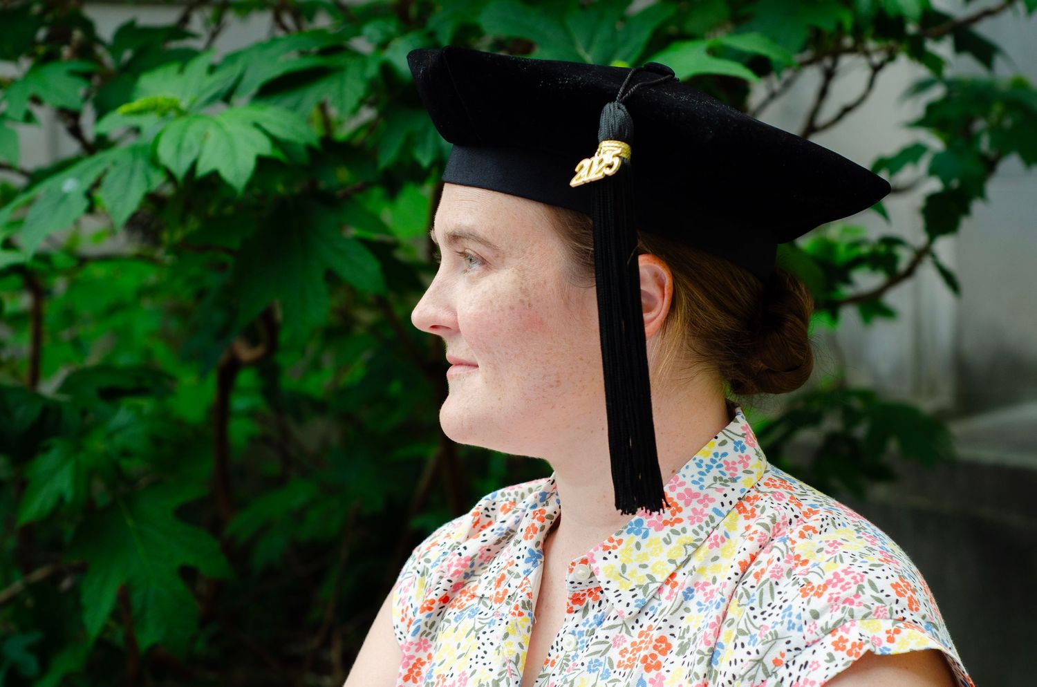 Graduate wearing academic cap and floral dress poses outdoors near greenery.