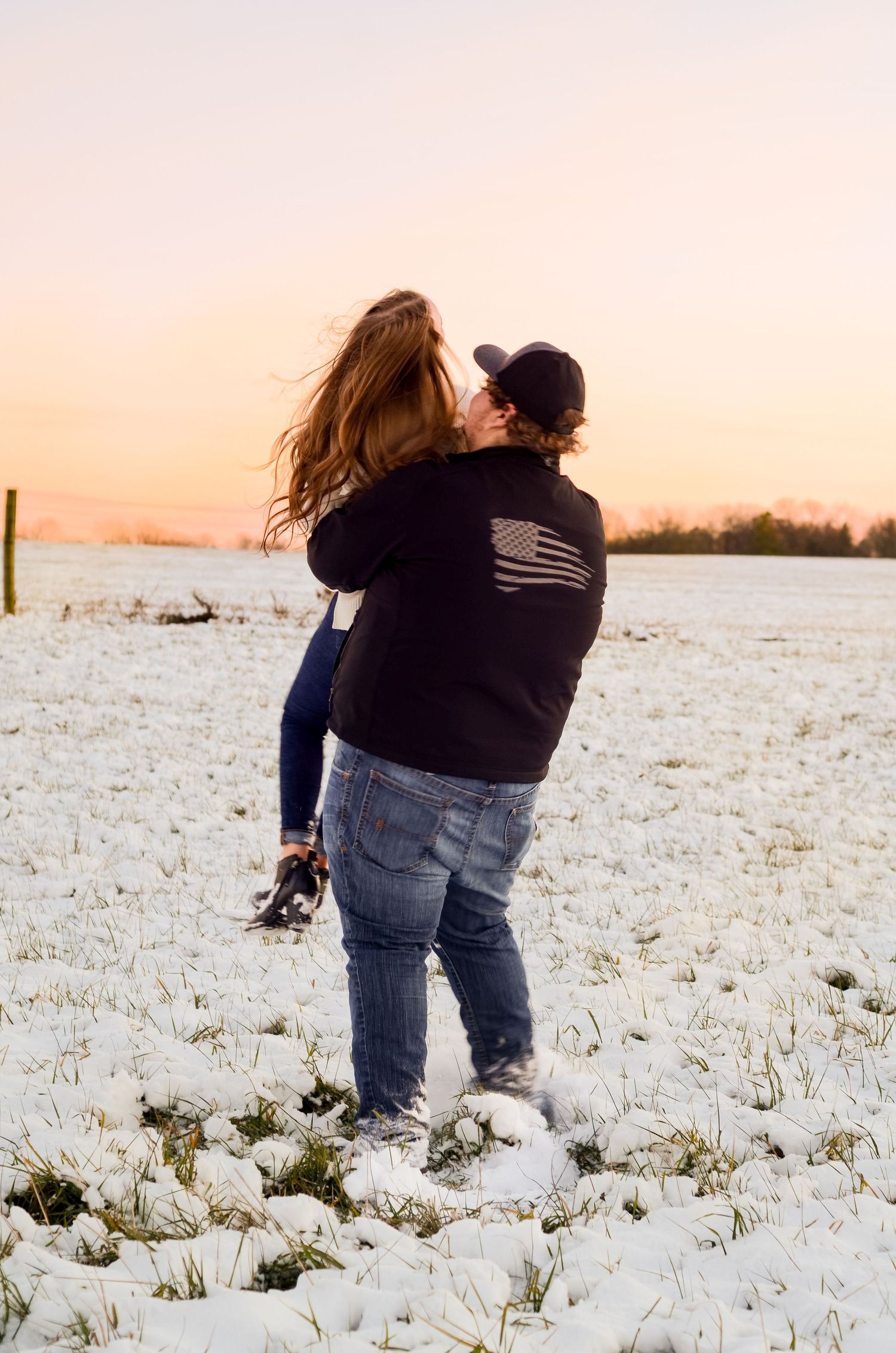 A couple embraces in a snowy field at sunset with one wearing a black sweater with flag design.
