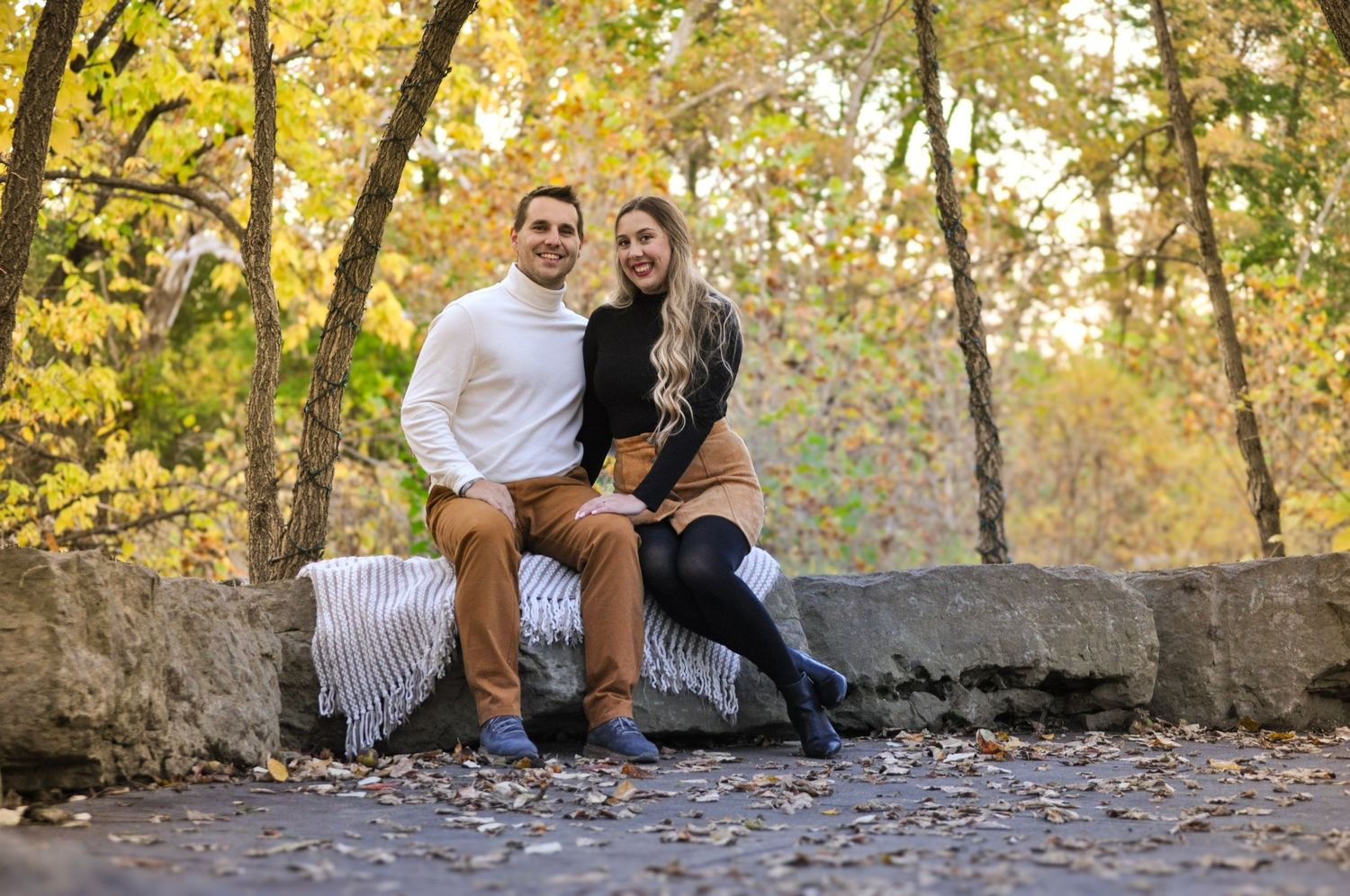 Couple sits together on stone wall surrounded by autumn foliage in park setting.
