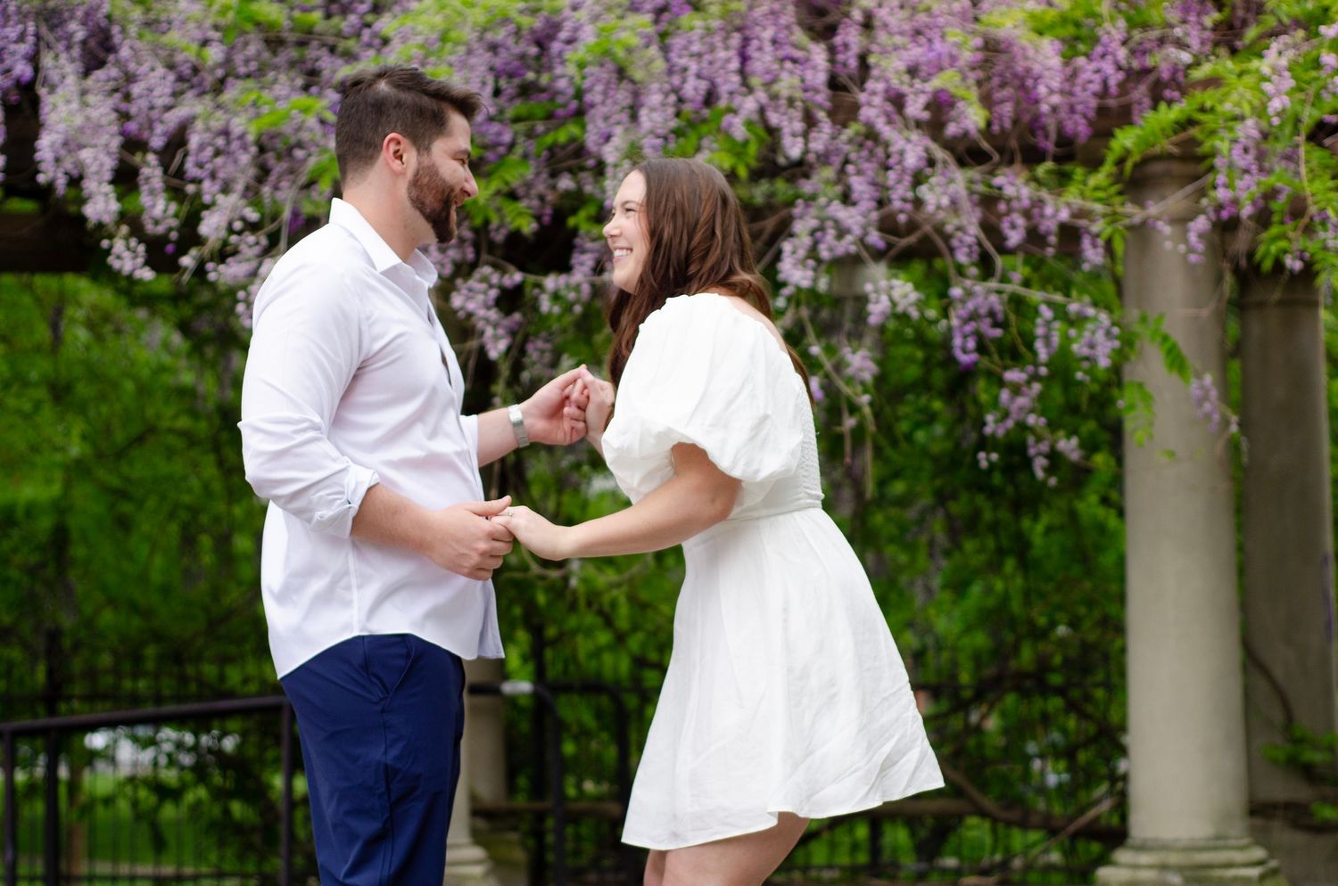 Couple shares romantic moments under blooming wisteria flowers during springtime engagement photo session. Wedding photos by KM Conceptions