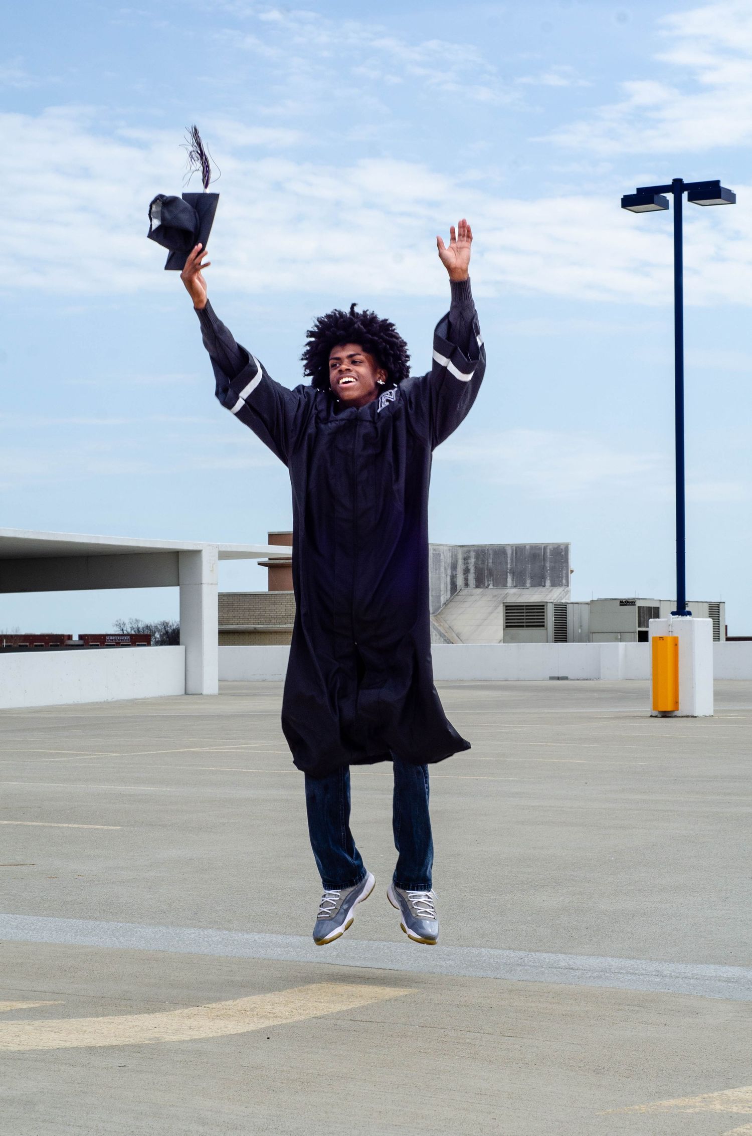A person in graduation attire jumps joyfully in a parking garage against a bright sky.