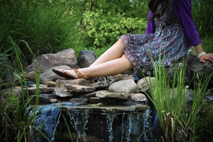A person's hands gently touch the flowing water of a natural stream or pond surrounded by rocks and greenery.