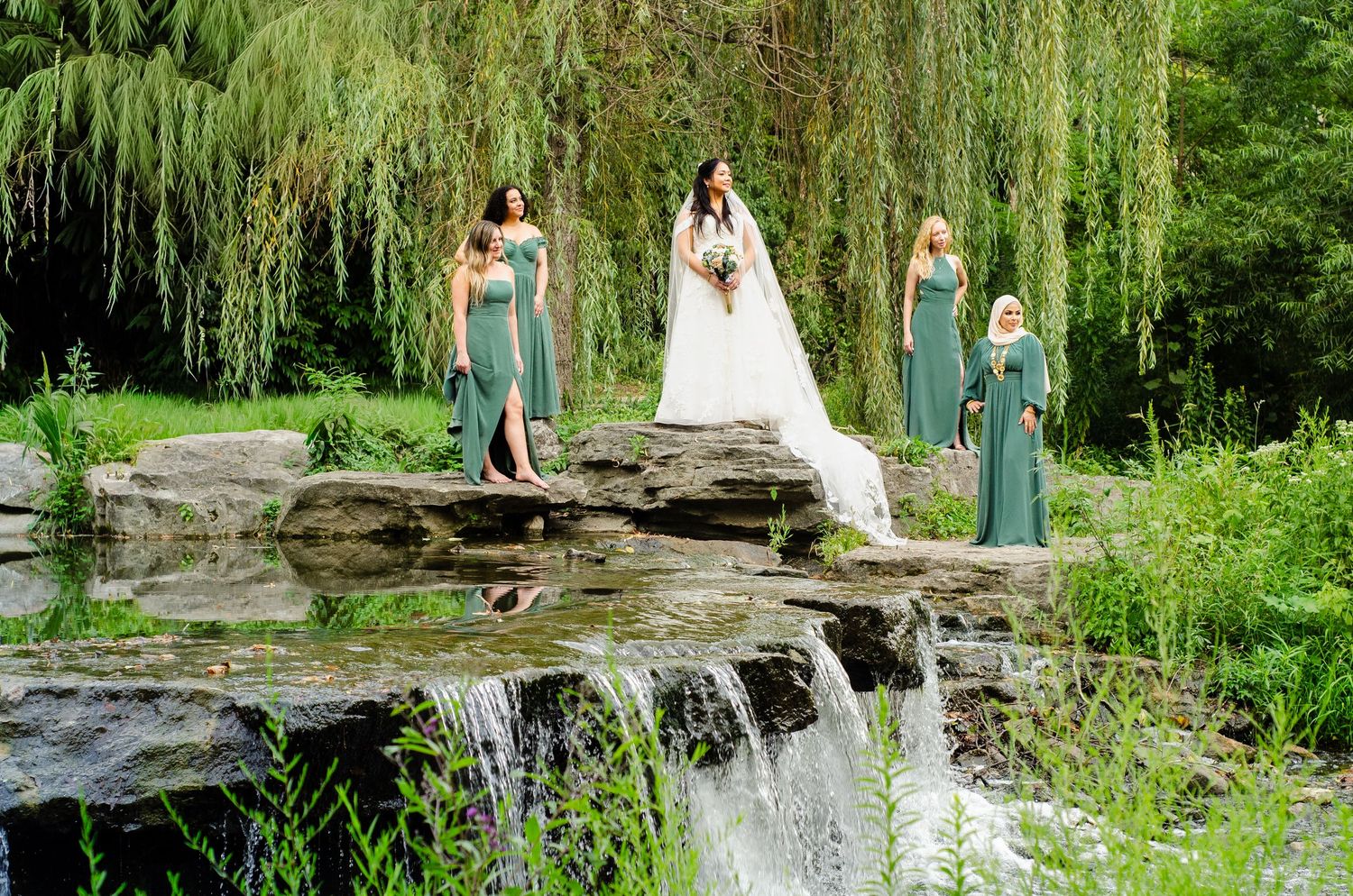 Wedding party poses on stone bridge near cascading waterfall surrounded by lush green willows and gardens.