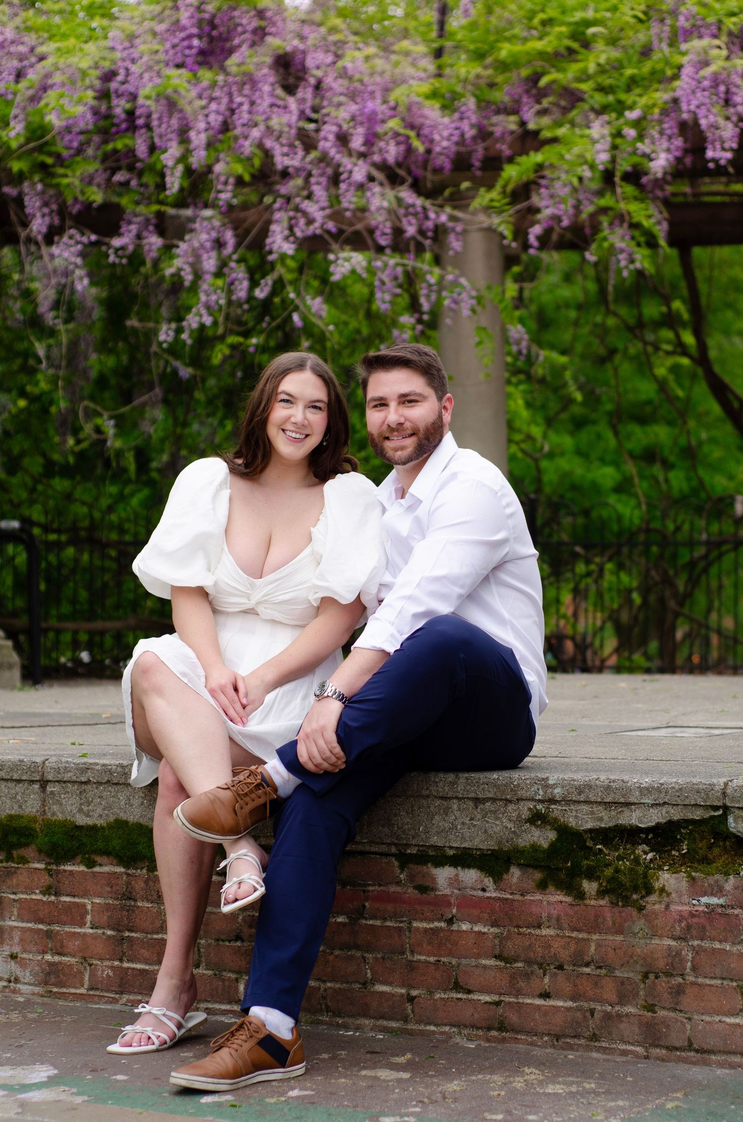 Couple sitting on brick wall under beautiful purple wisteria blooms.