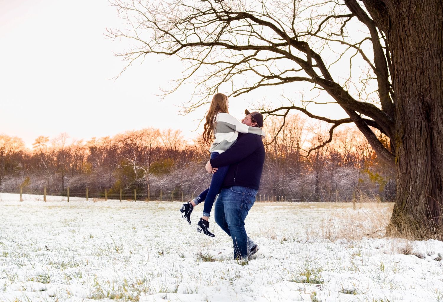 A person in winter clothing jumps playfully in deep snow near a bare tree at sunset.