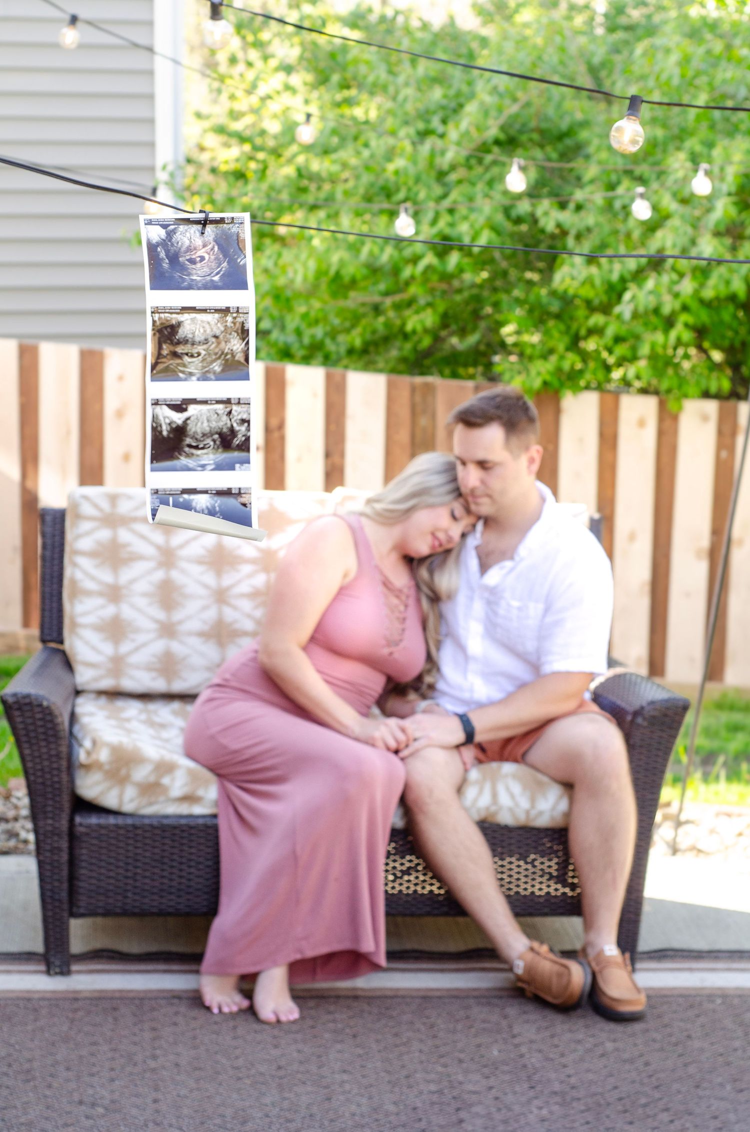A couple sits together on outdoor patio furniture with string lights and photos hanging in the background.