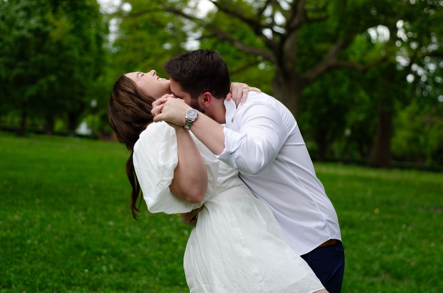 Playful couple in white attire embracing in a lush green outdoor setting.
