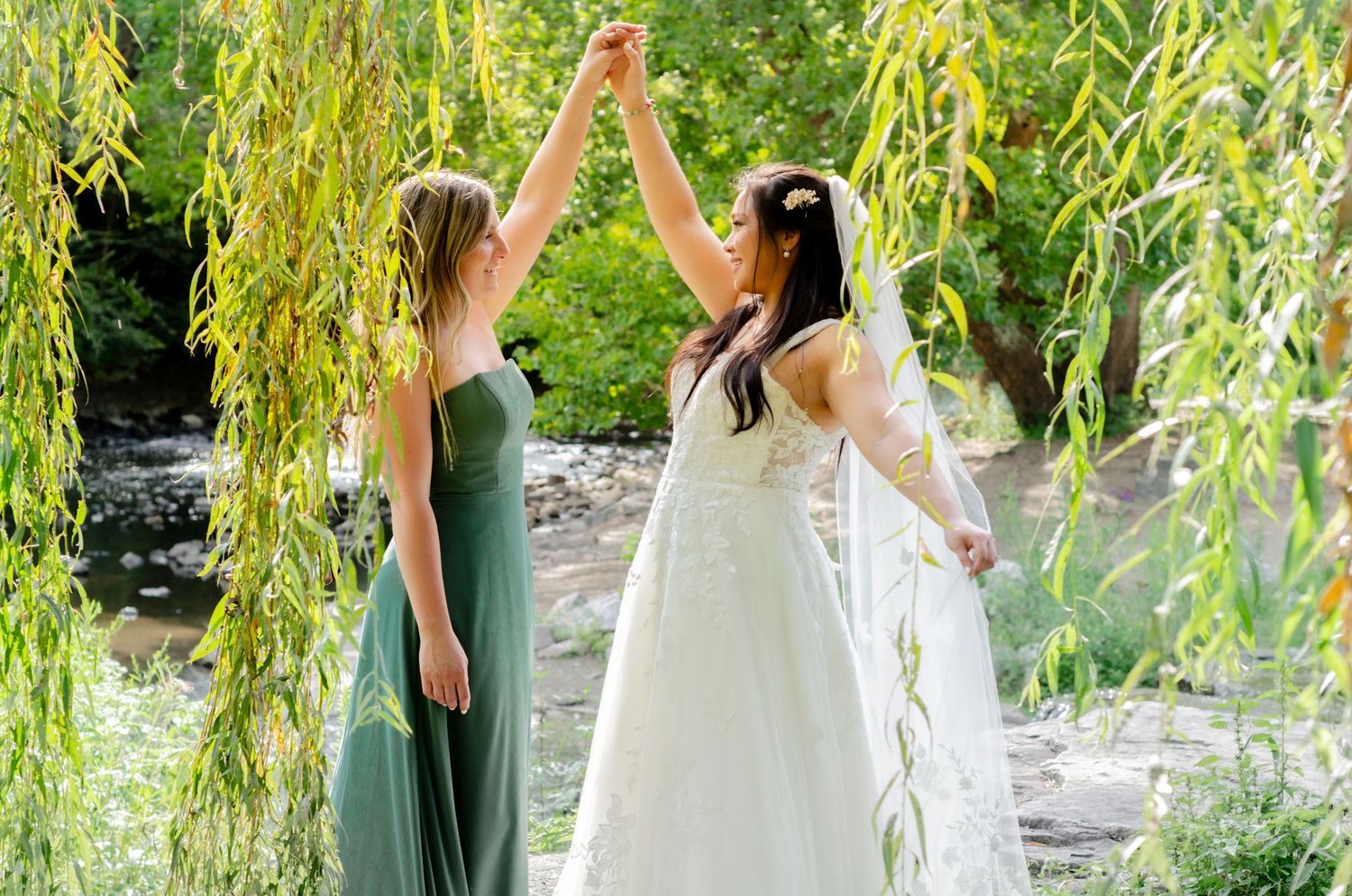 Wedding party in green dresses poses playfully on a fallen log by a pond under drooping willow trees.