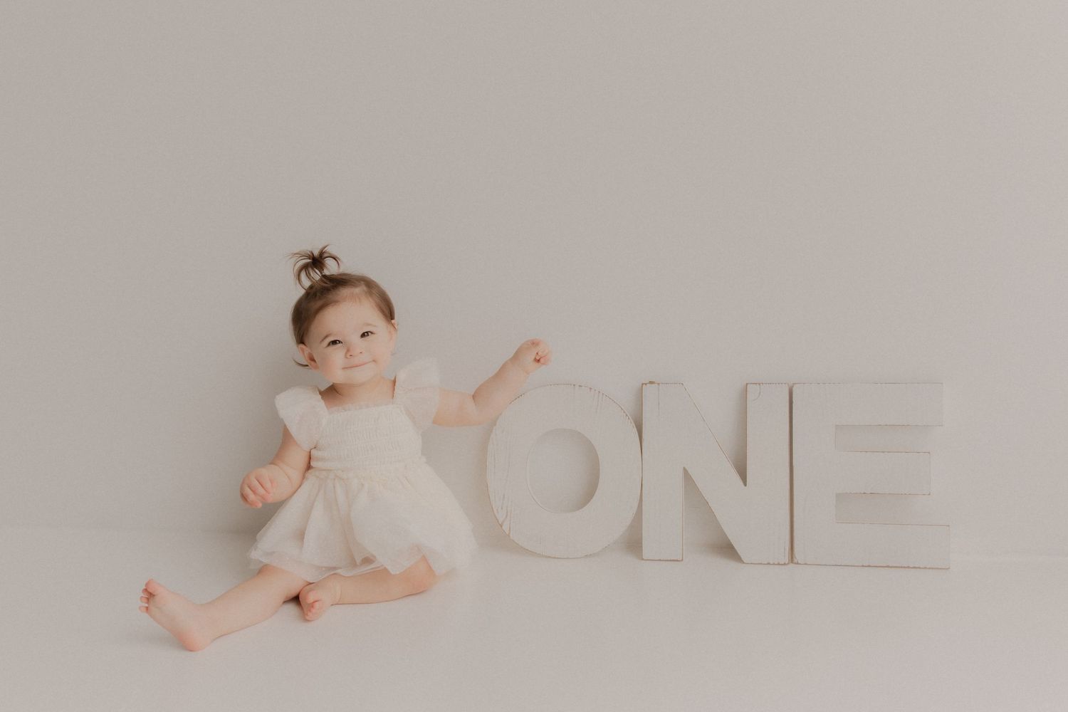 Baby wearing white dress sits next to large white ONE letters against neutral background for first birthday photo.