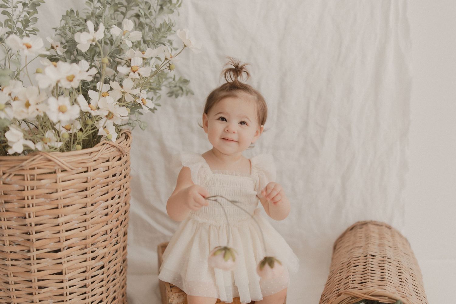 A small child in a white dress sits beside a wicker basket with floral decorations against a light backdrop.