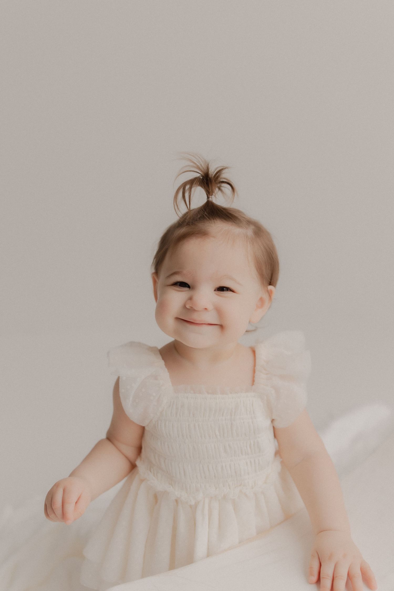 A small child wearing a white ruffled dress smiles brightly against a neutral beige background.