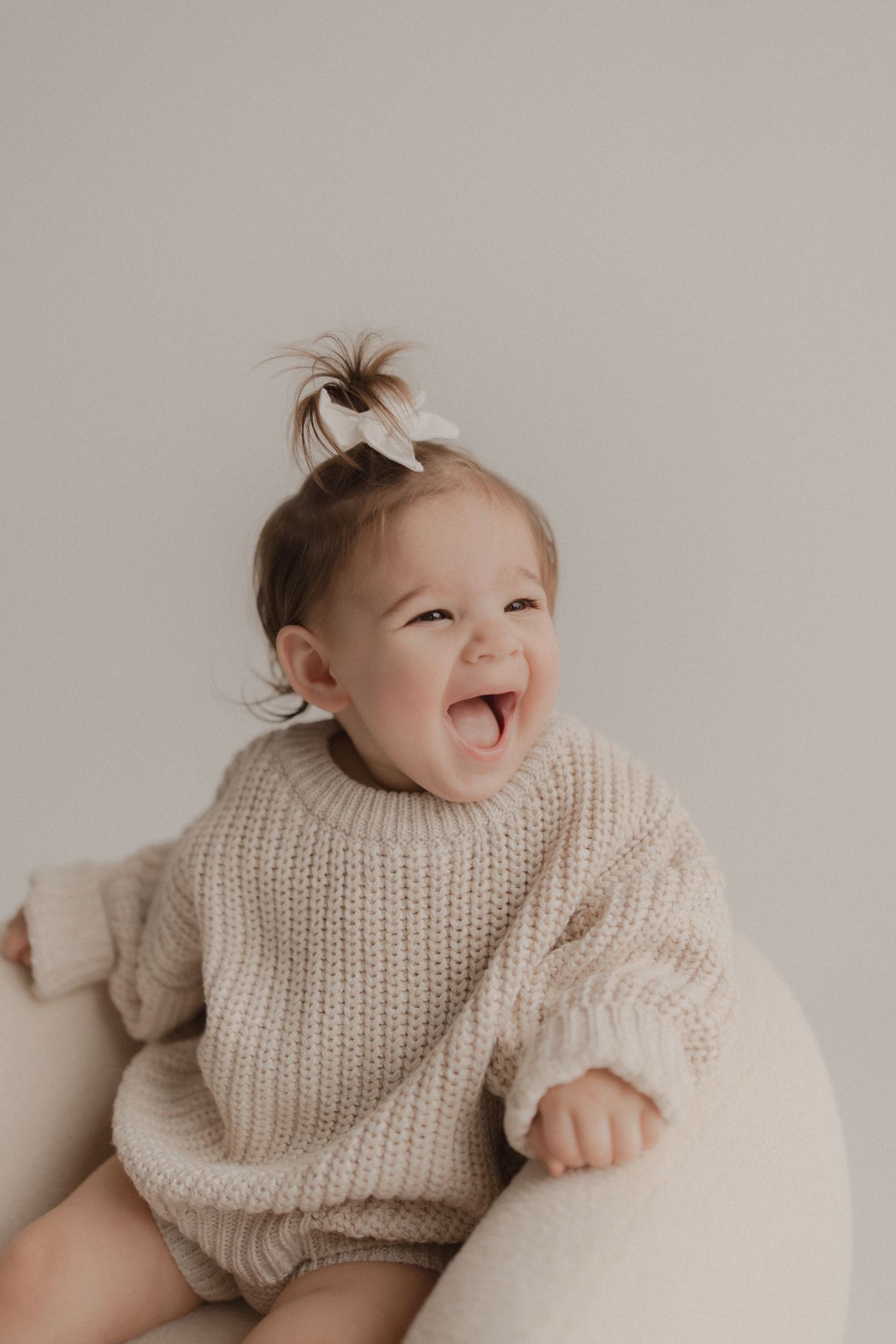 Baby in cozy cream knit sweater laughs joyfully while sitting on light colored chair.