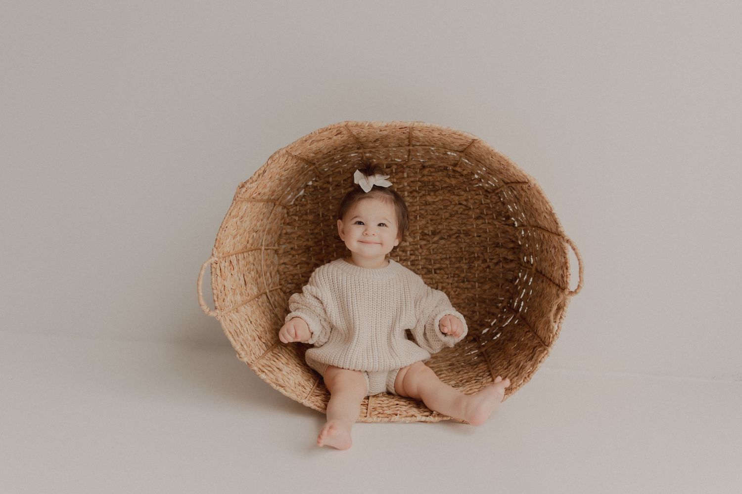 A toddler sits nestled inside a large round woven basket against a neutral background.