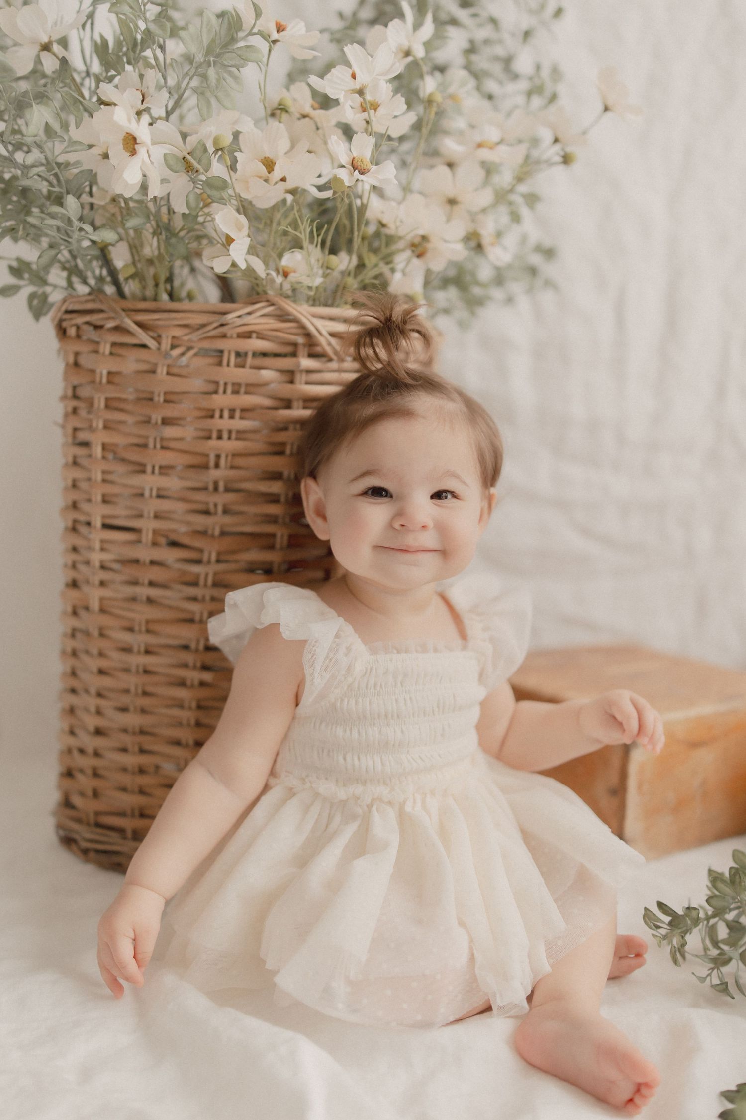 A small child wearing a white eyelet dress sits on a soft white surface next to a woven basket filled with flowers.