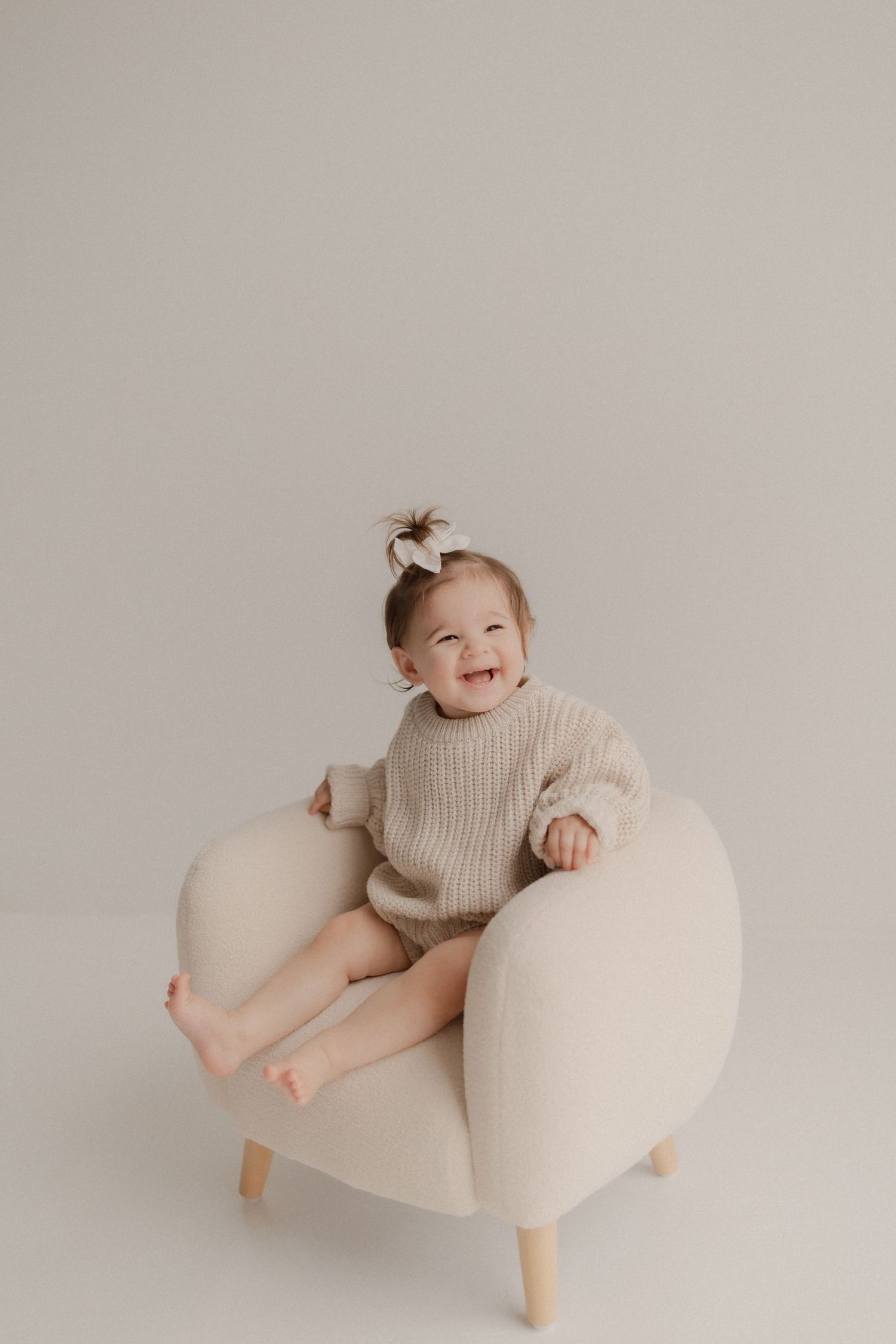 A small child wearing a knit sweater sits happily on a cream colored modern accent chair against a neutral background.
