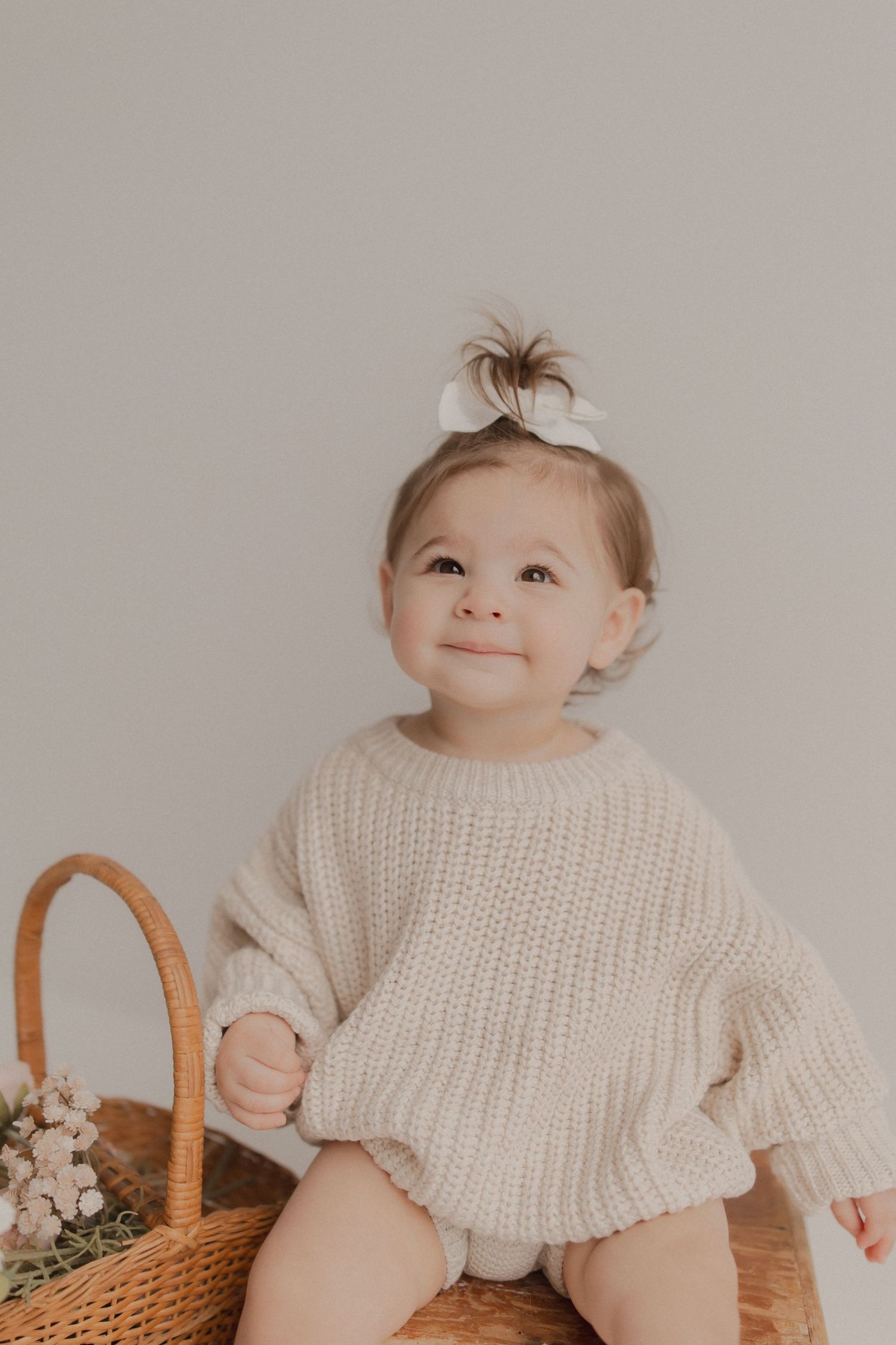 Sweet baby in cream knit sweater sitting beside woven basket with flowers against neutral background.