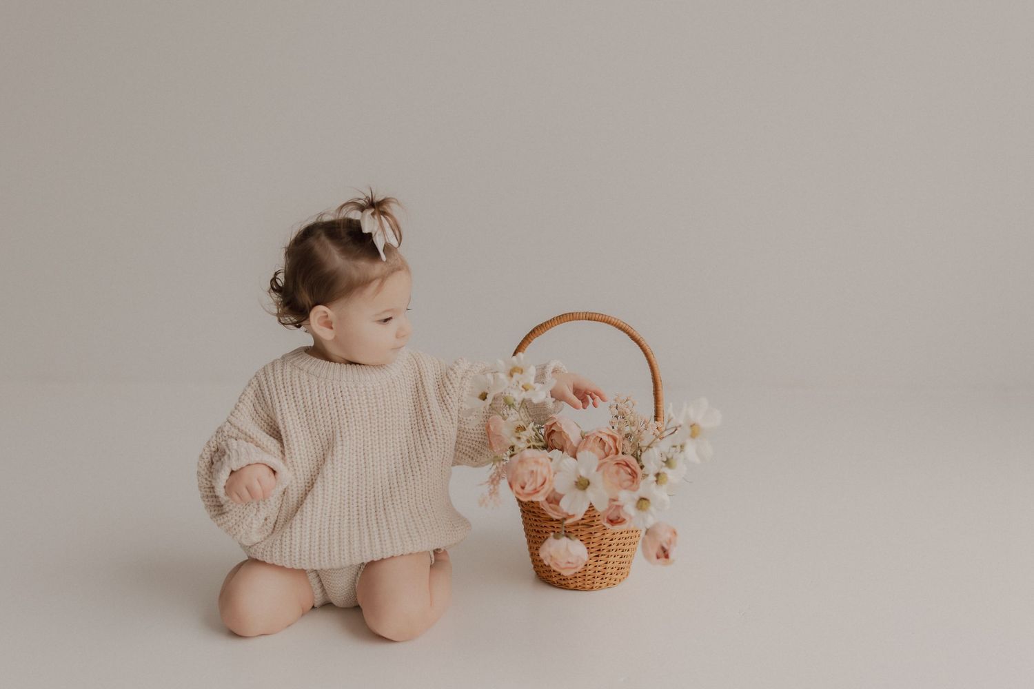 A child in a cream sweater sits beside a wicker basket filled with pink and white flowers on a neutral backdrop.