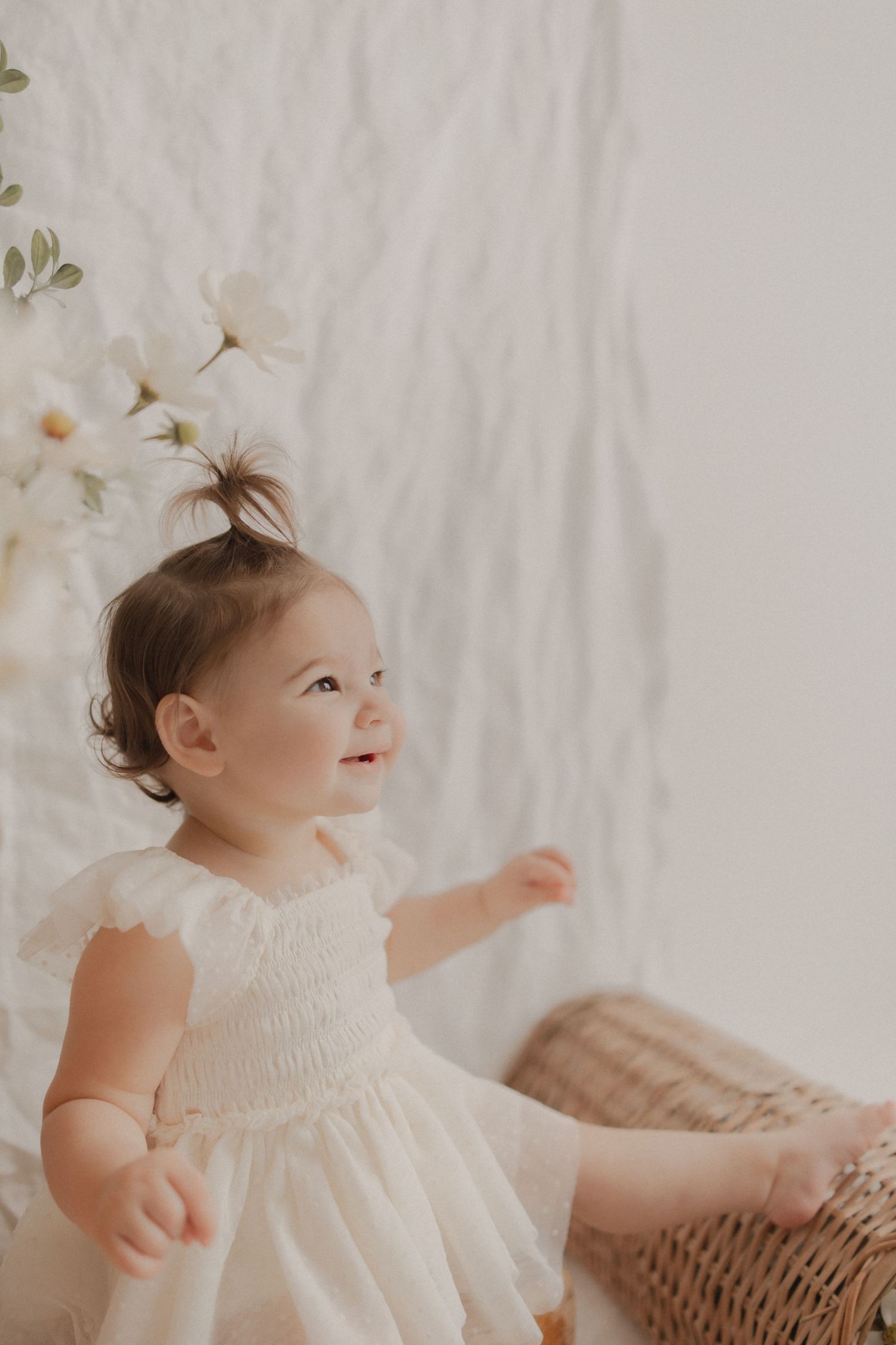 A small child wearing a delicate white smocked dress smiles while standing near white flowers and wicker furniture.