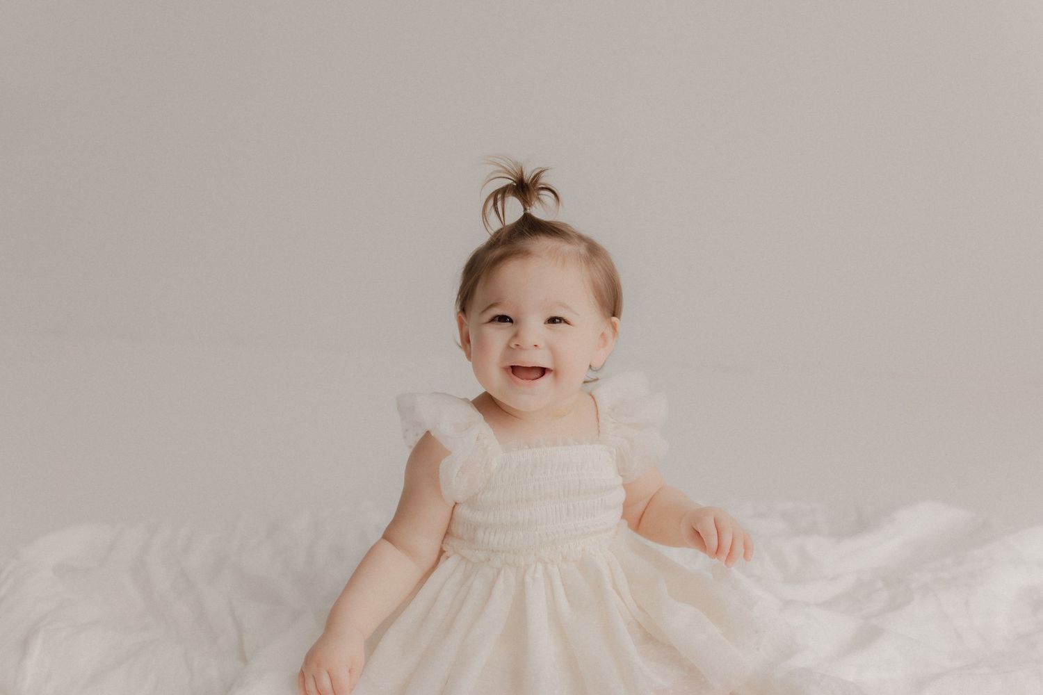Small baby wearing a white dress smiles while sitting on soft white bedding.