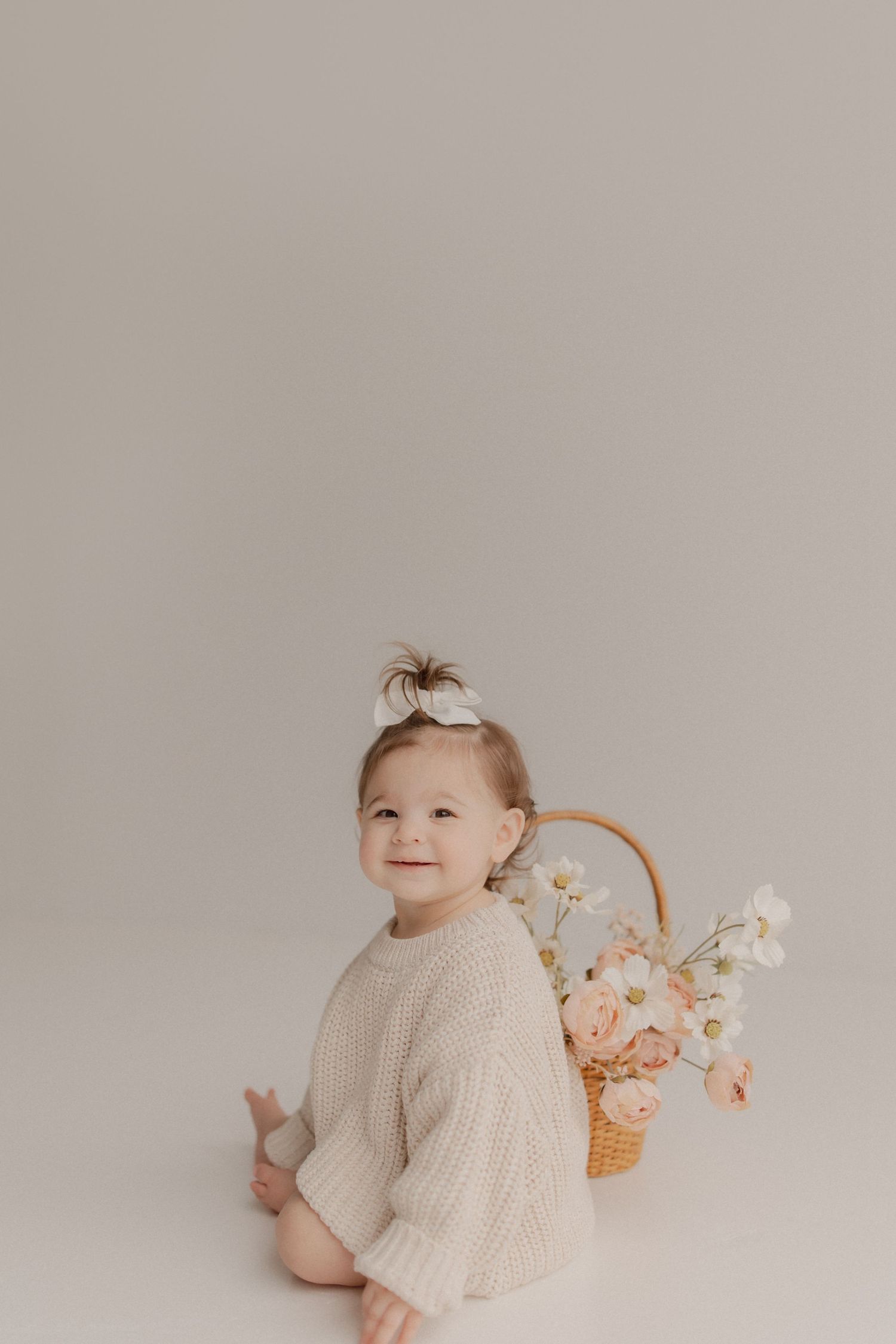 Smiling baby in cream sweater poses beside wicker basket filled with pink flowers against neutral backdrop.