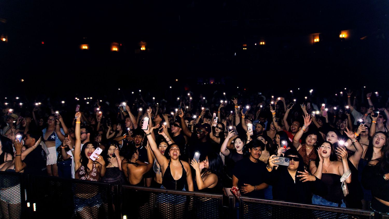 Concert crowd with raised hands silhouetted against bright stage lights during a nighttime performance.
