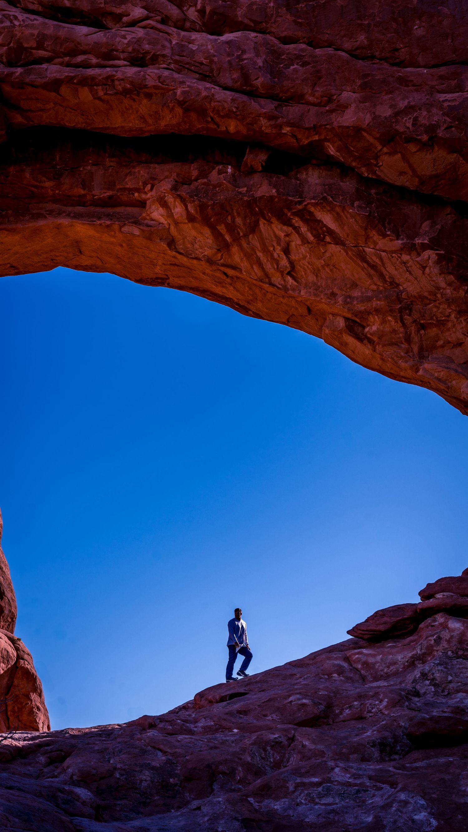 A silhouetted figure stands beneath a natural stone arch against bright blue desert sky.