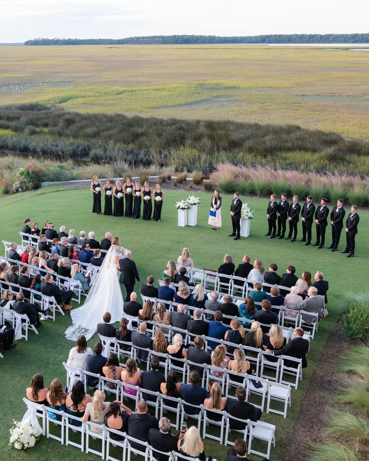 Aerial view of an outdoor wedding ceremony on a green lawn overlooking coastal marshlands and water at sunset.