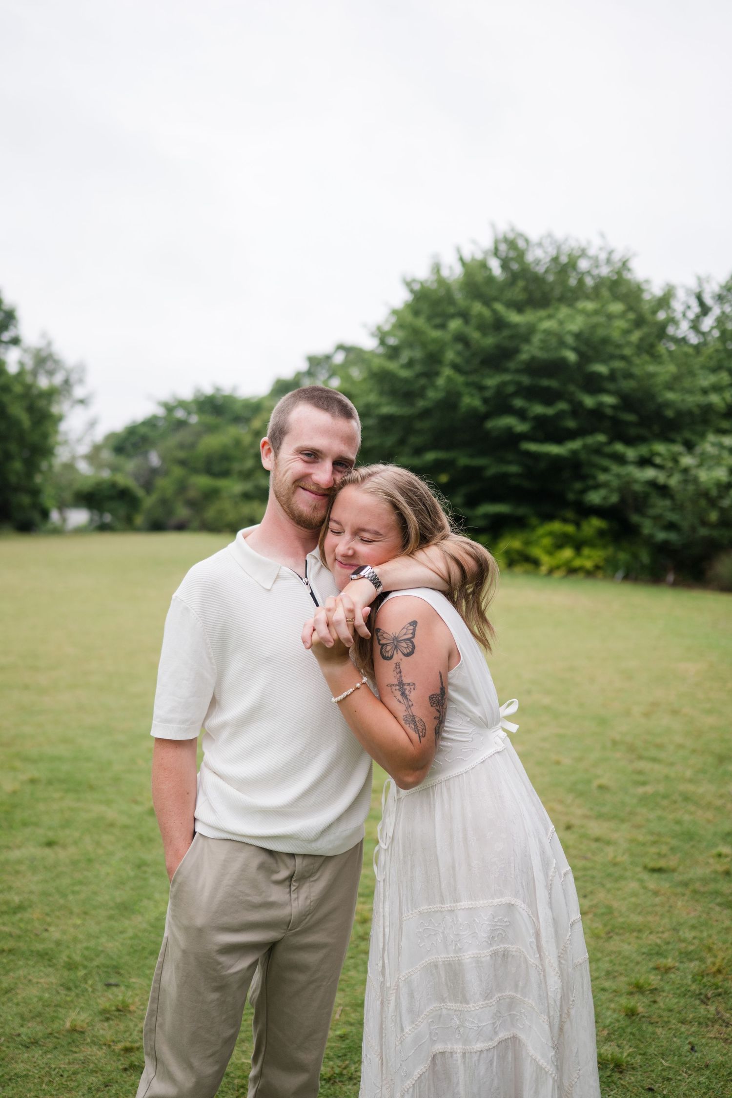 A couple shares joyful candid moments together in a grassy field while wearing white and beige casual outfits.
