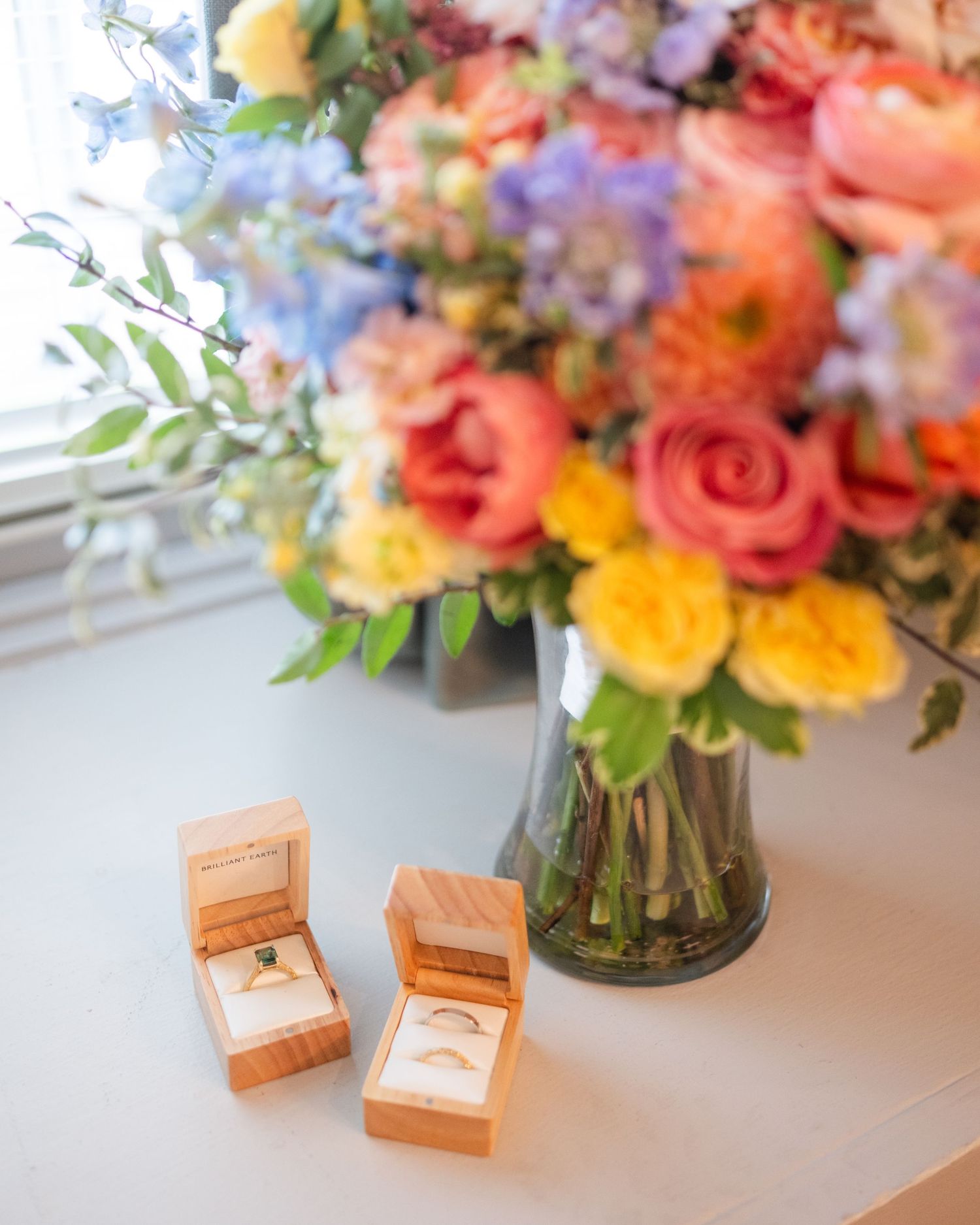 Two wedding rings in wooden boxes beside a colorful floral bouquet with pink, yellow, and blue blooms in a glass vase.