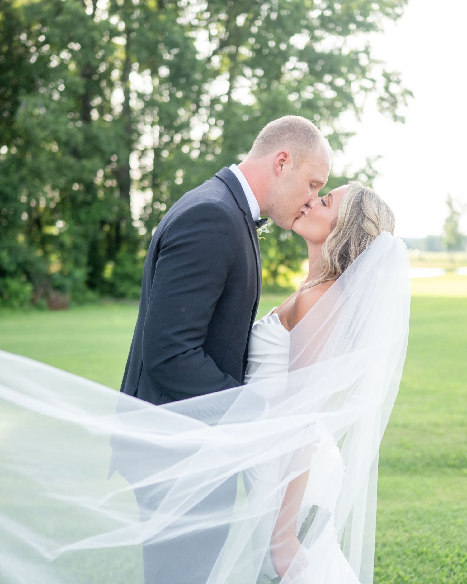 A romantic wedding couple embraces in a lush green garden setting as the bride's veil flows gracefully in the breeze.