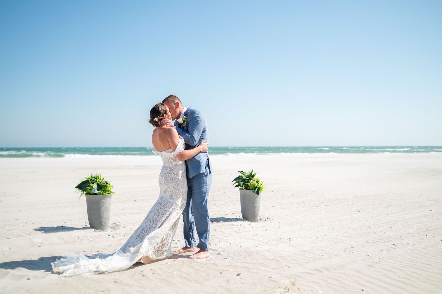 A couple embraces on a pristine white sand beach with potted plants and the ocean in the background.
