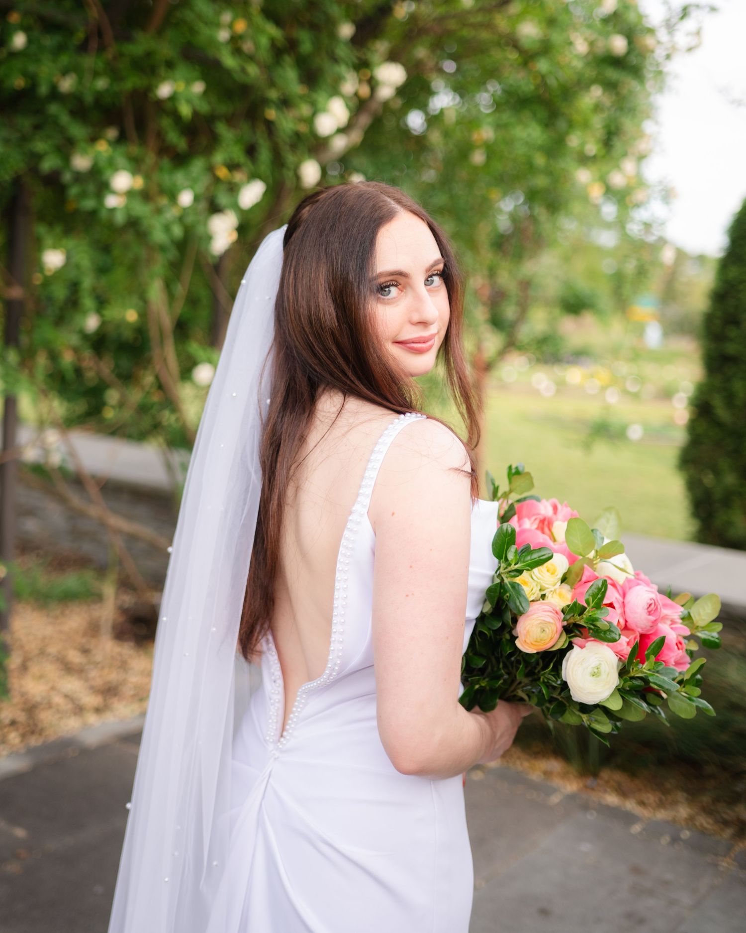 A bride wearing a white gown and veil poses with a pink and peach rose bouquet in a garden setting.