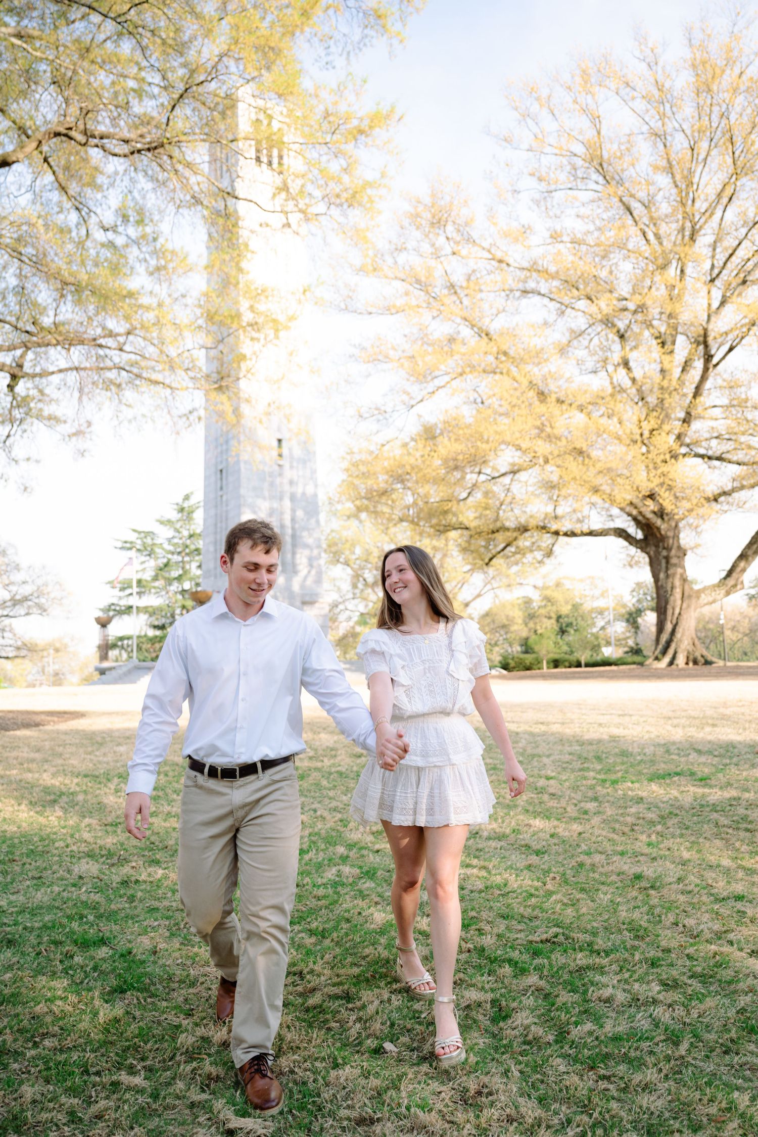 Happy couple holding hands and walking outdoors among golden trees during a sunny engagement photo session.