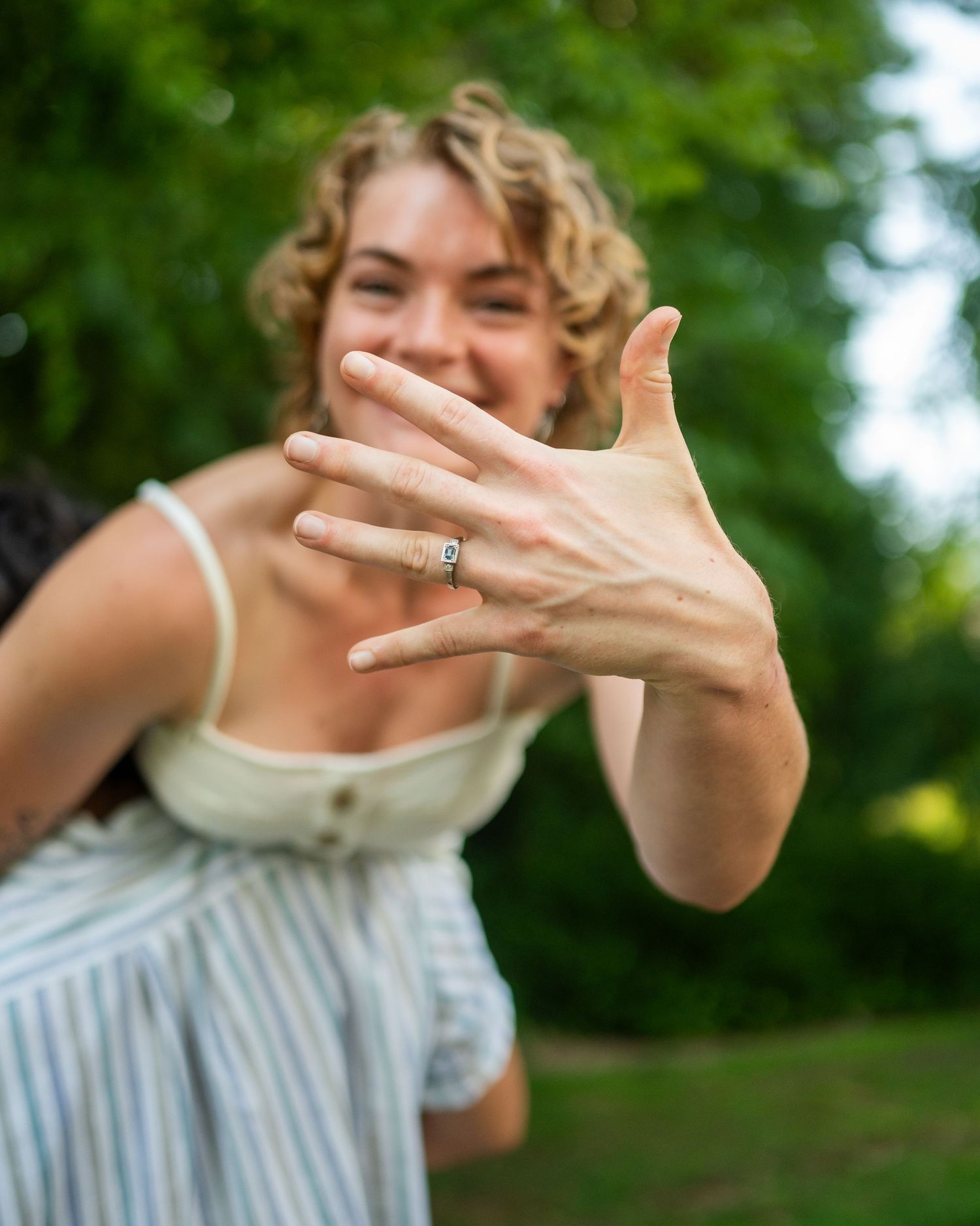 A hand displaying an engagement ring outdoors with a blurred green background.