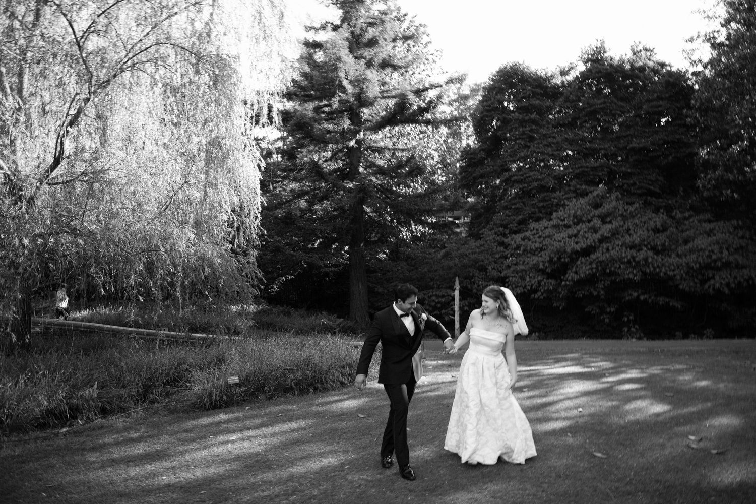 Black and white photo of a bride and groom holding hands walking along a tree-lined path on their wedding day.
