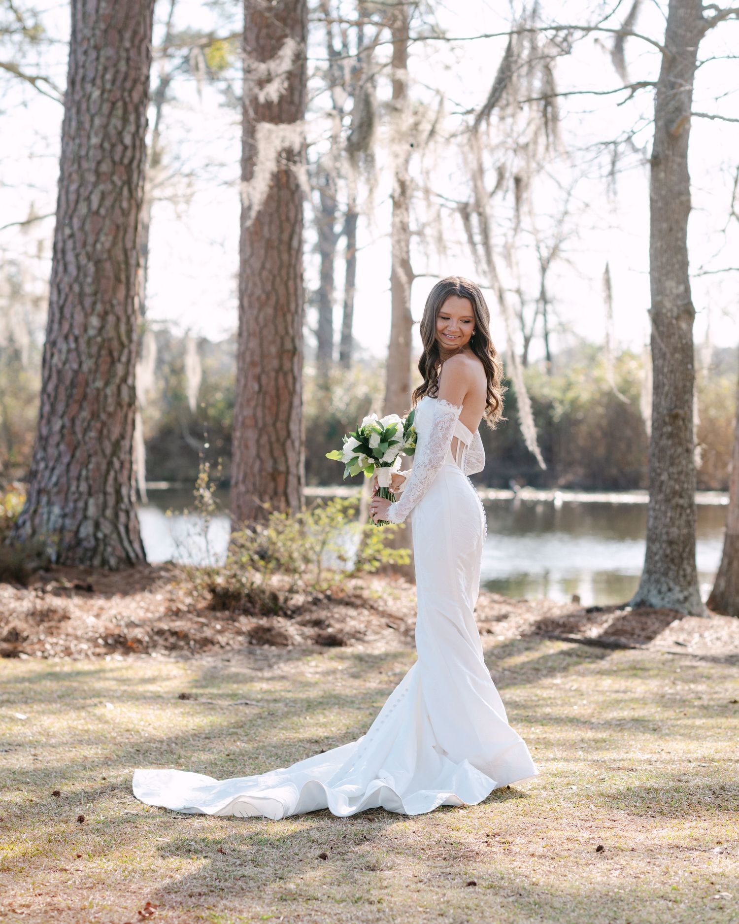 Bride in white mermaid gown with train holds bouquet outdoors near pond surrounded by tall trees.