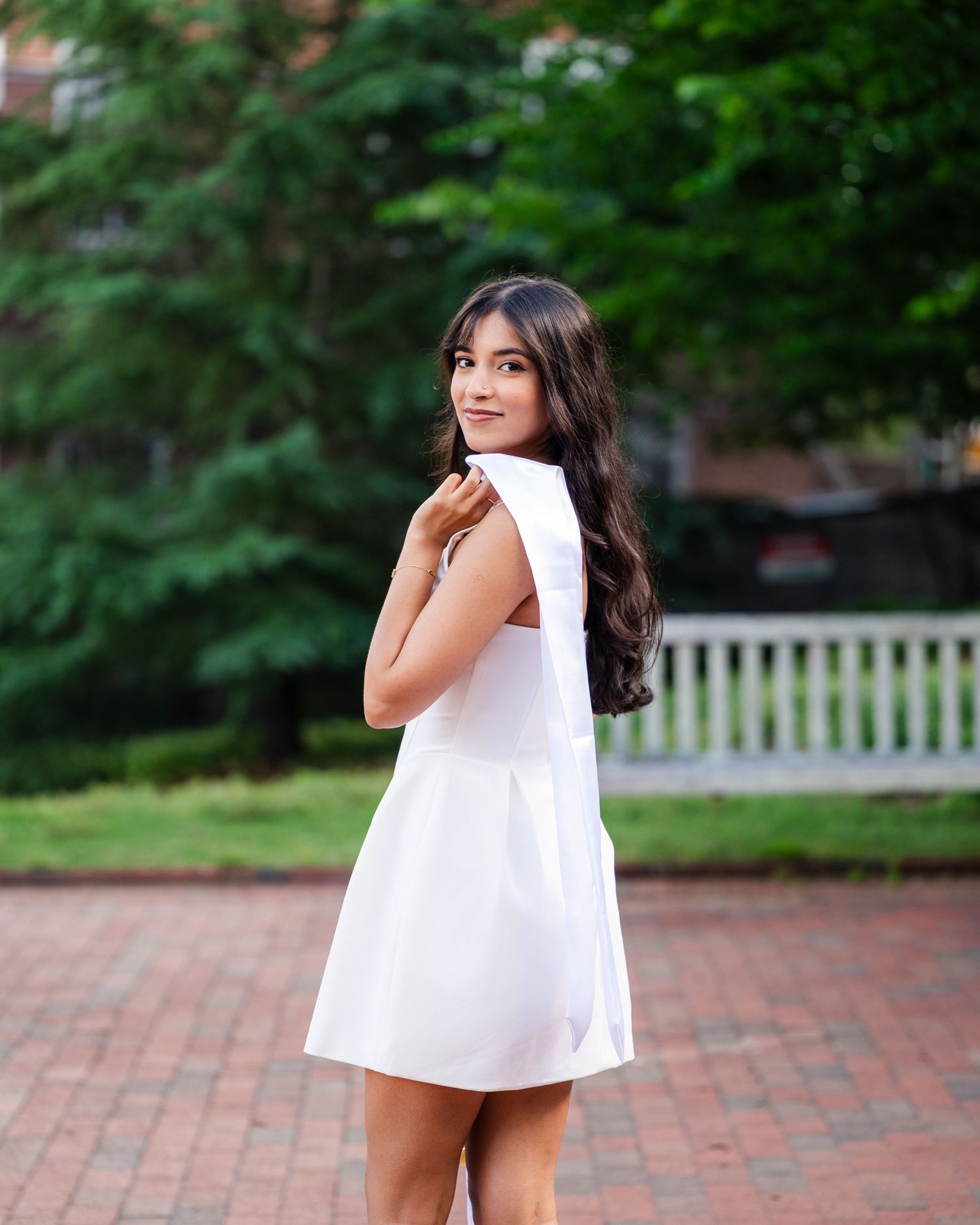 A person in a white sleeveless dress poses playfully outdoors on a brick walkway surrounded by greenery.