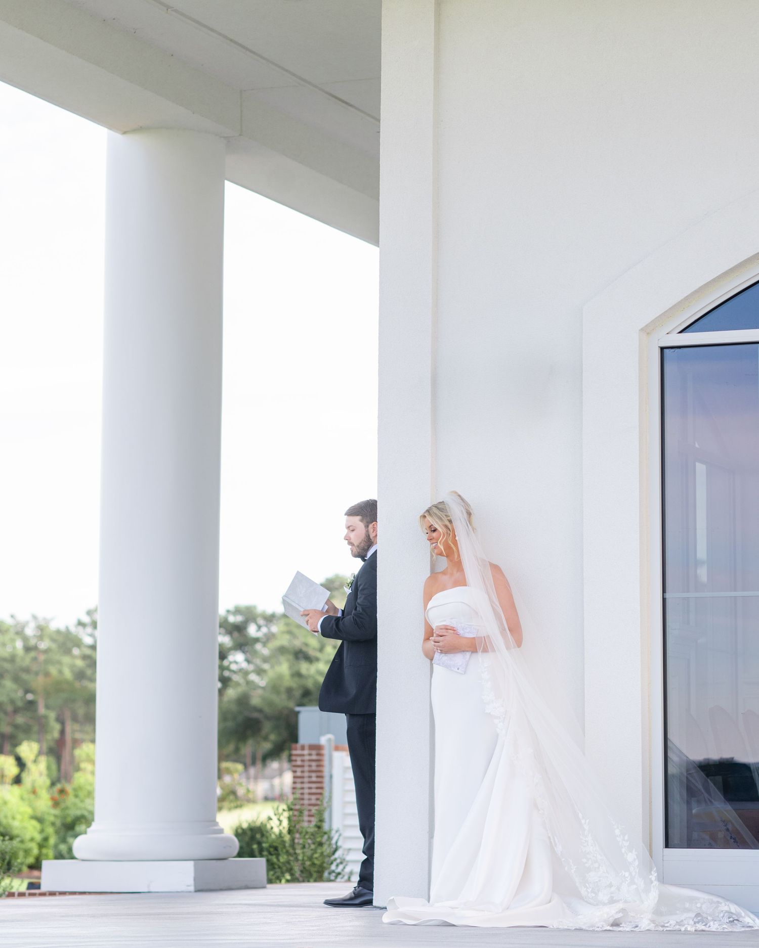 Bride in flowing white dress poses elegantly between white columns on a bright modern porch with greenery in background.