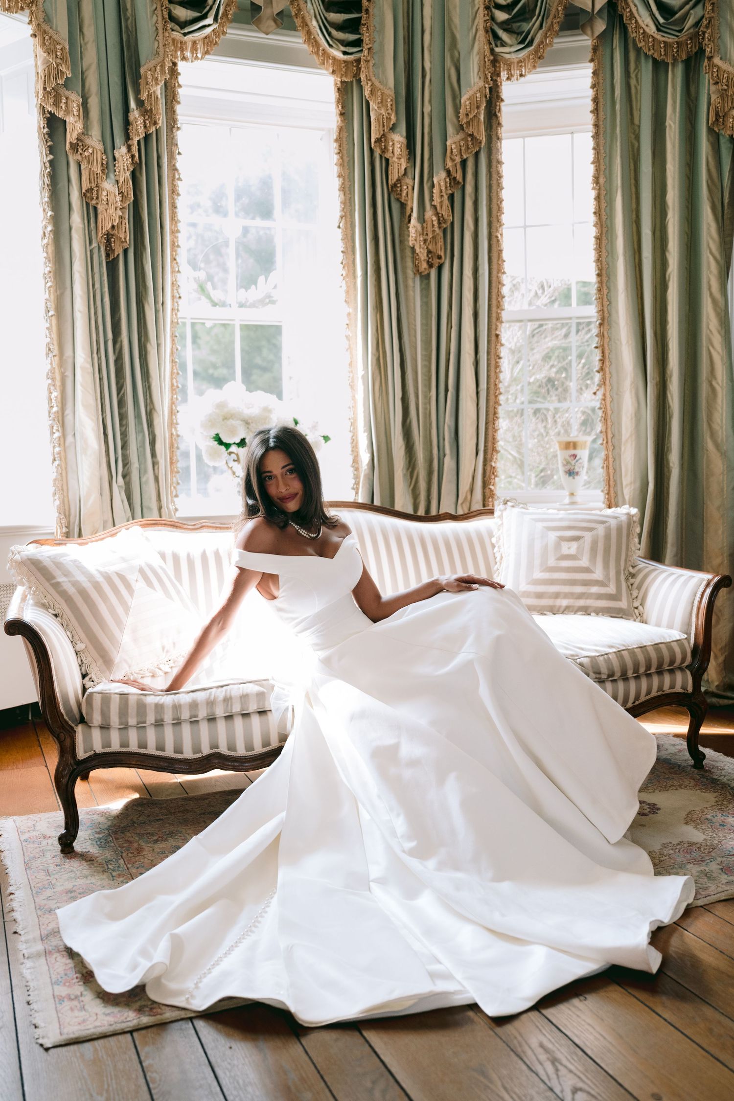 Bride in white off-shoulder gown sitting on striped sofa with elegant green drapes and hardwood floors.