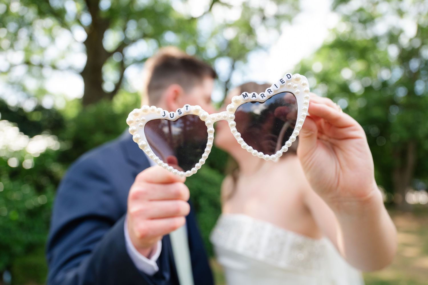 Couple kissing at wedding holding heart-shaped pearl sunglasses on sticks in outdoor garden setting.