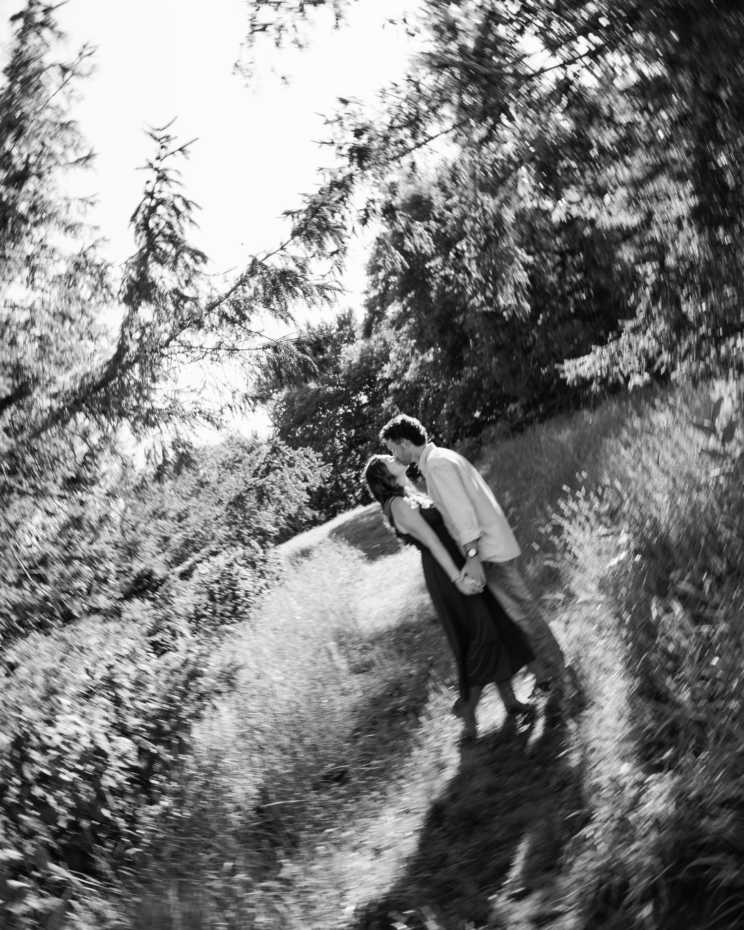Black and white photo of a couple kissing on a sunlit forest path surrounded by lush trees.