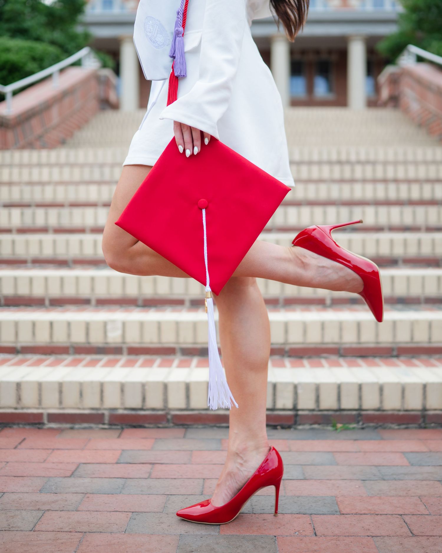 Graduate in white dress holds red diploma folder and poses on brick steps wearing matching red high heels.